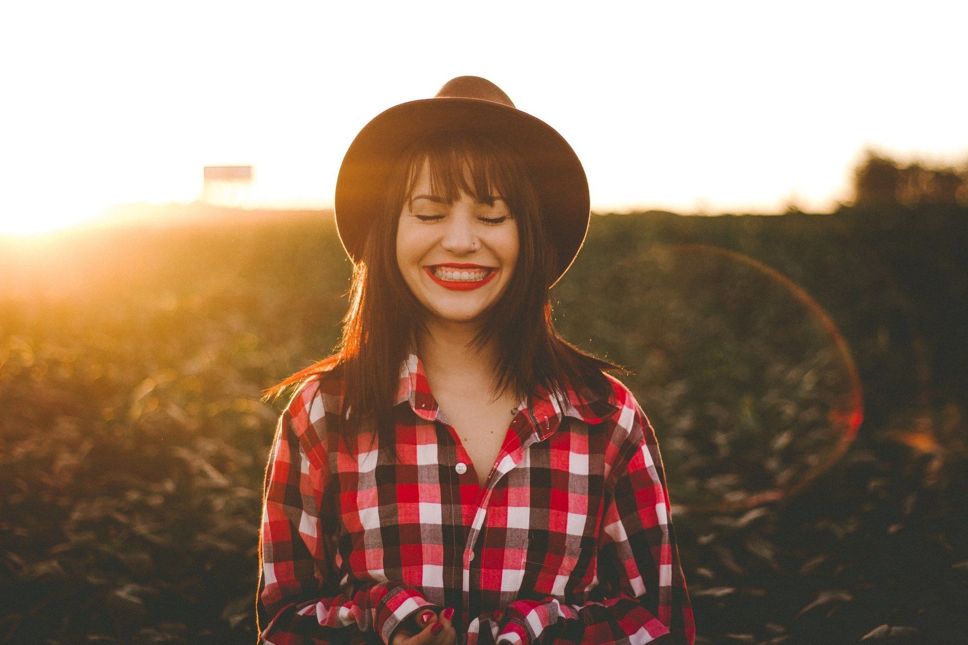 A woman wearing a hat and a plaid shirt is smiling in a field.