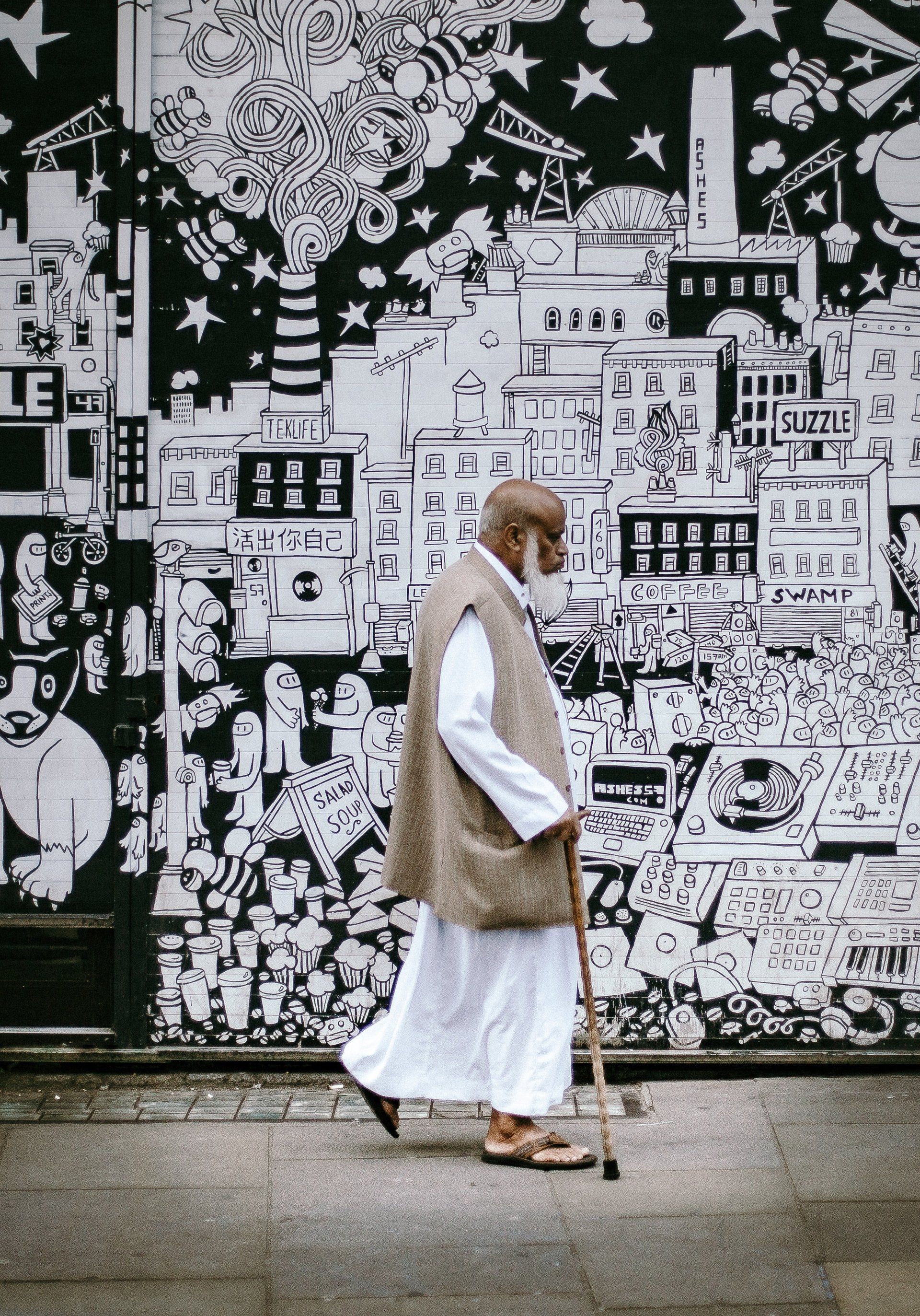 A man with a cane is walking in front of a wall with a drawing of a city on it