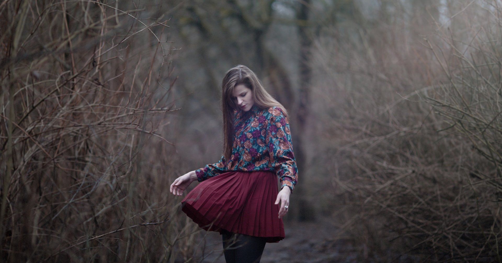 A woman in a red pleated skirt is standing in the woods.