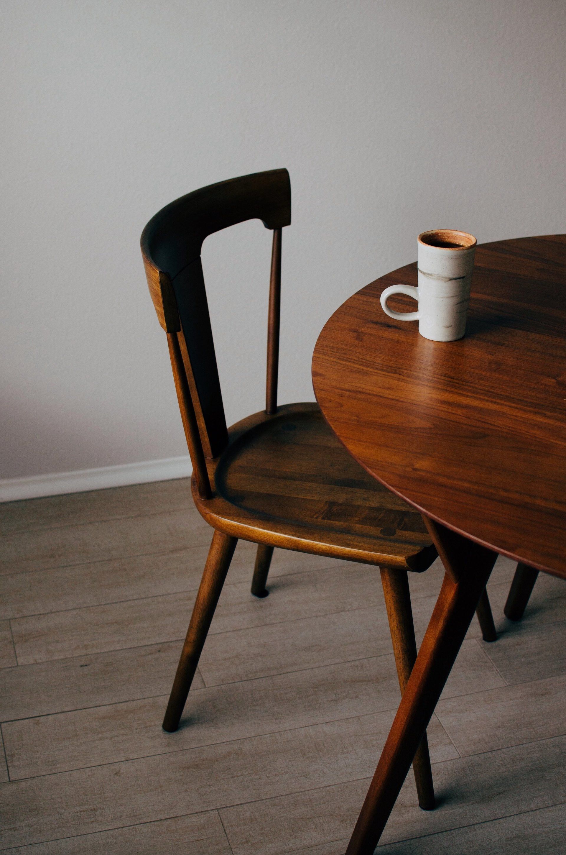 A wooden chair is sitting next to a wooden table with a cup of coffee on it.