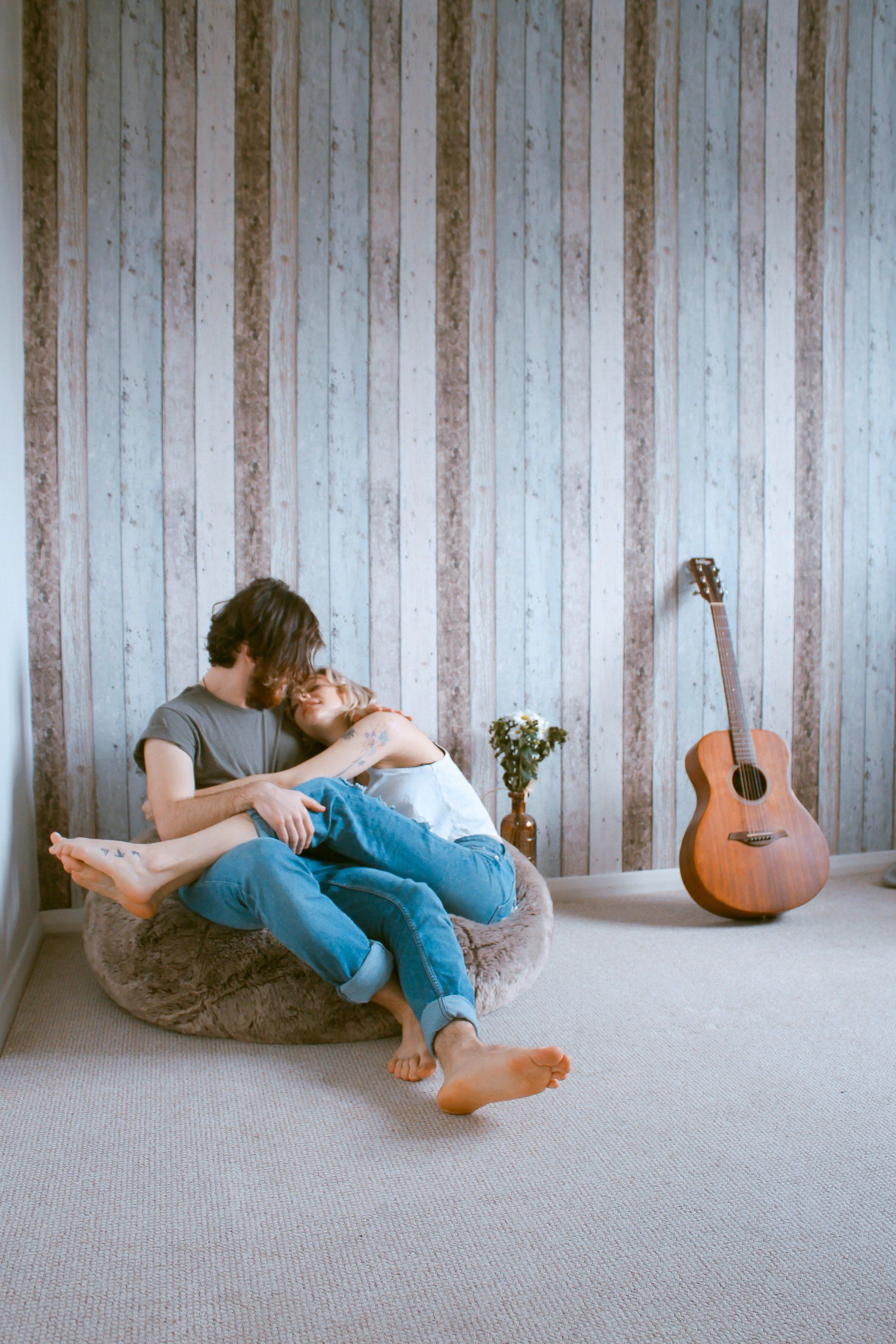 A man and a woman are sitting on a bean bag chair hugging each other in a room with a guitar.