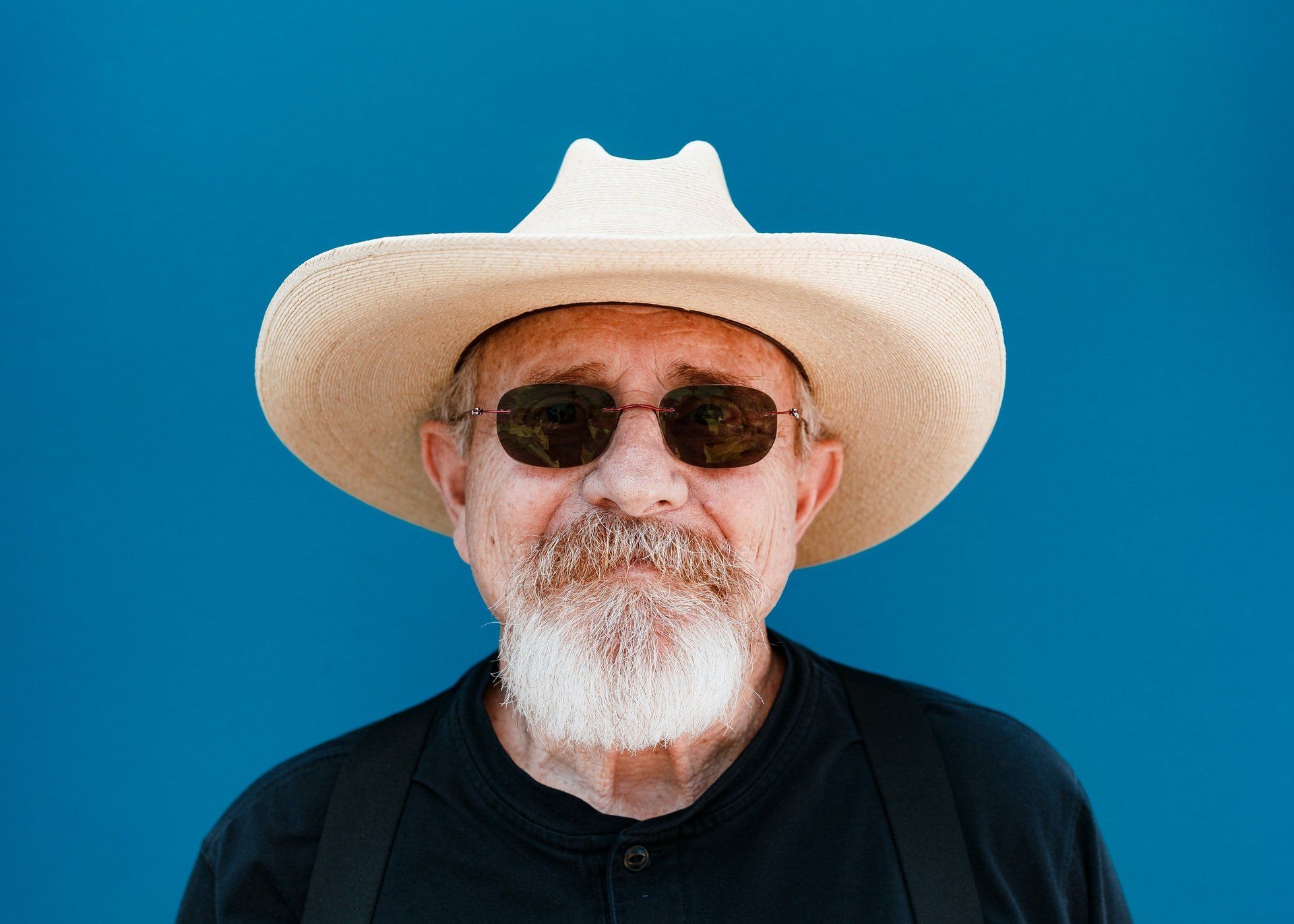 Man with white beard, sunglasses, and straw hat against a blue background.