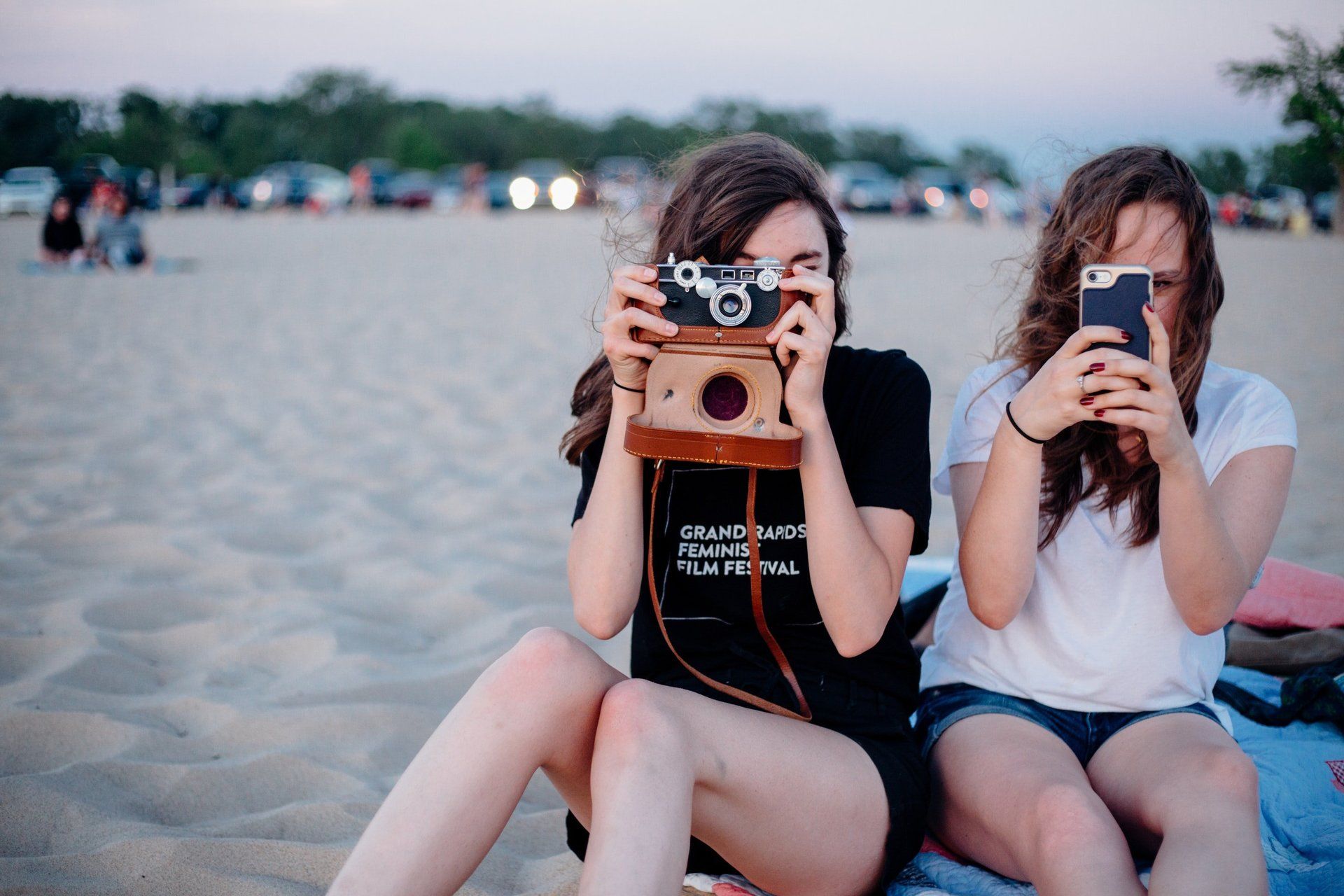 Two girls are sitting on the beach taking pictures with their cell phones.