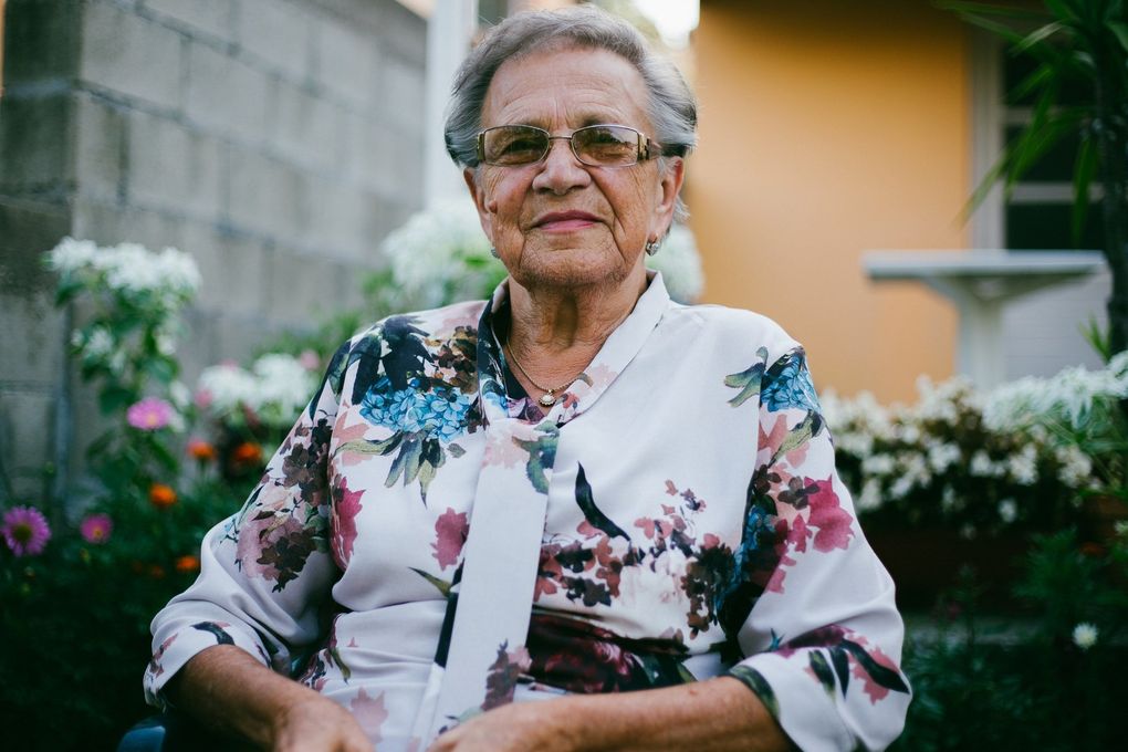 An elderly woman wearing glasses and a floral shirt is sitting in a garden.