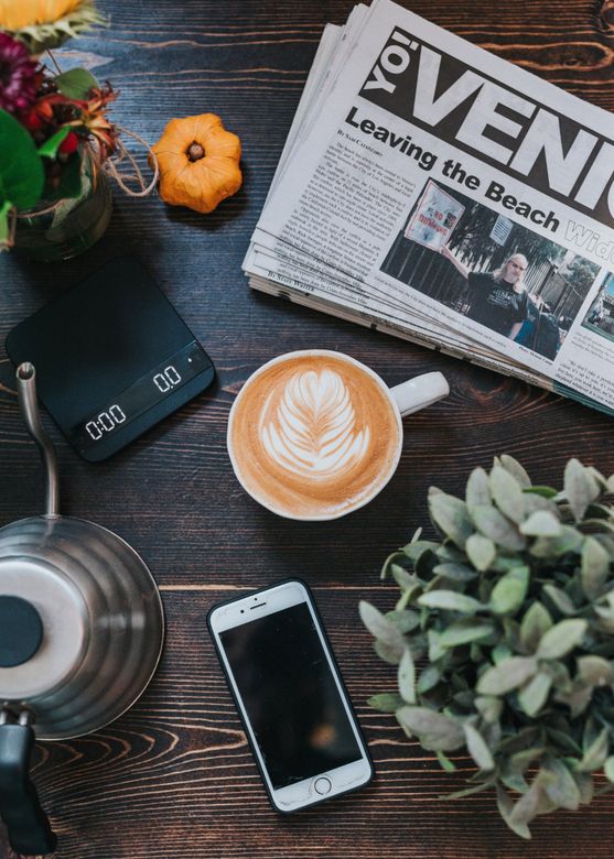 A cup of coffee , a newspaper , a phone , and a scale are on a wooden table.