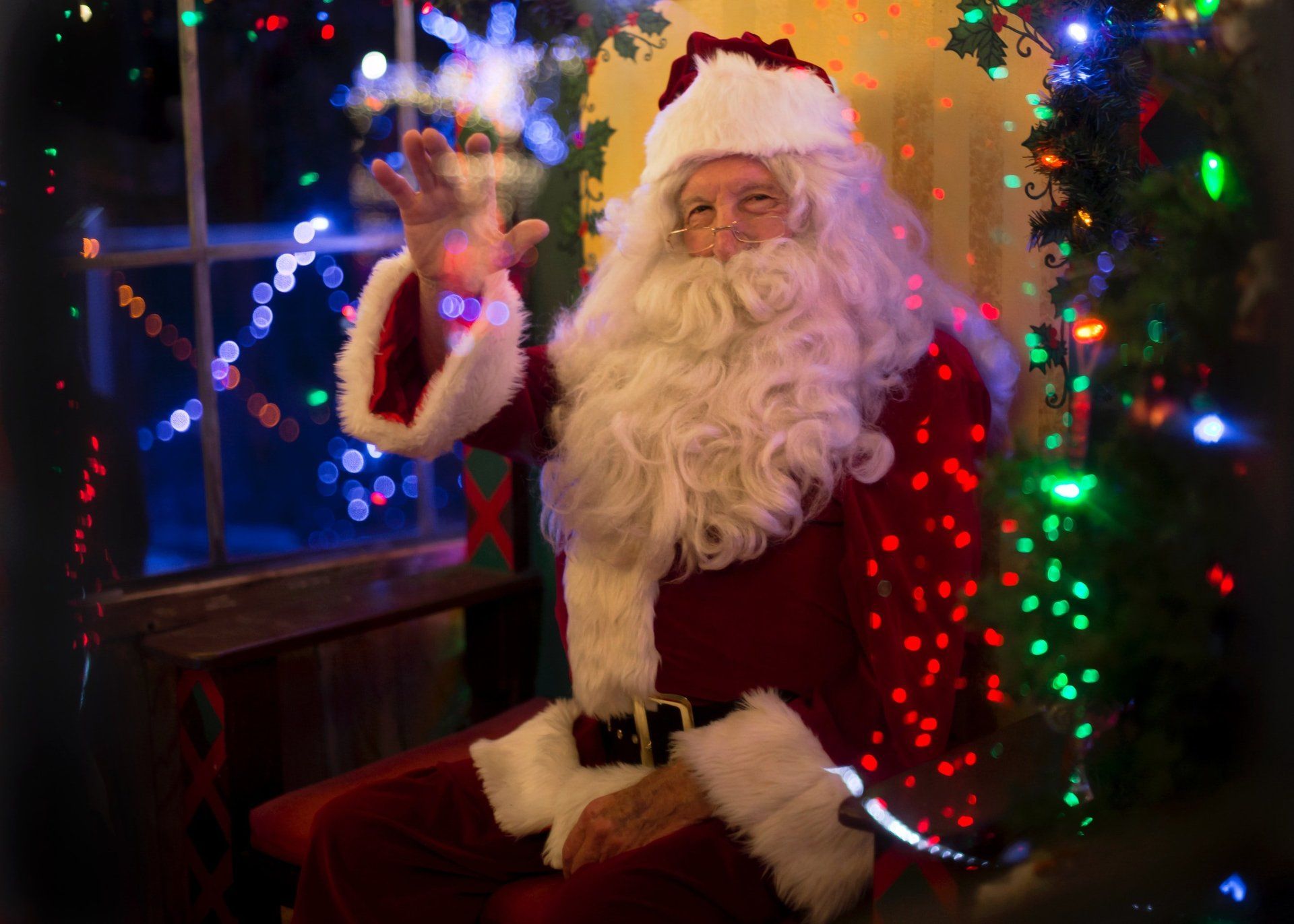 Santa Claus, in red suit, waving and smiling, sits by decorated Christmas tree and window.