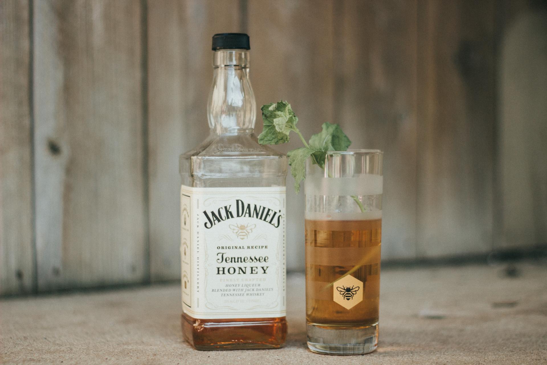 A bottle of Jack Daniel's Tennessee Honey whiskey next to a filled glass with a sprig of greenery.