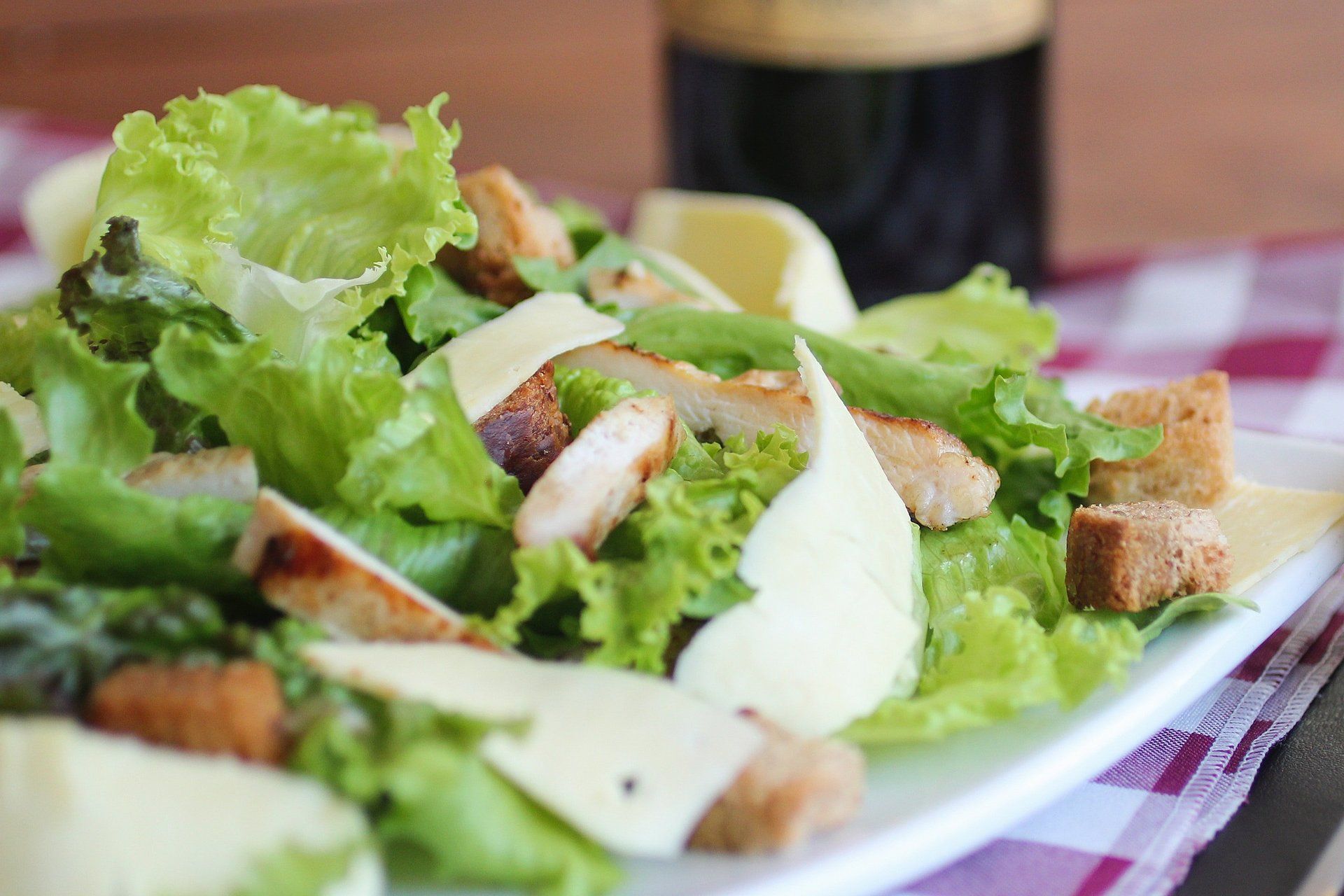 Chicken Caesar salad with lettuce, croutons, and cheese on a white plate; a dark bottle sits in the background.