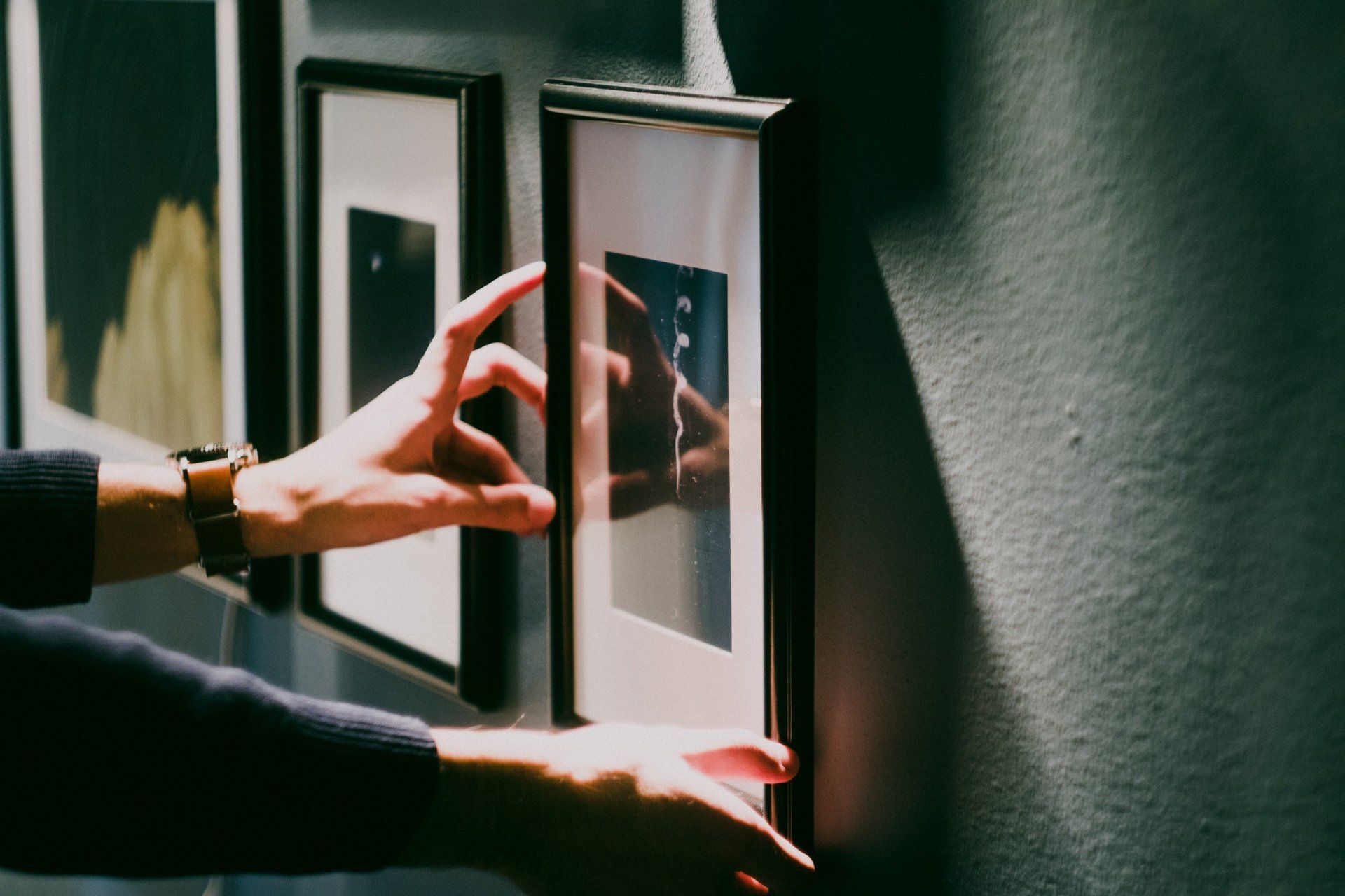 Hands adjusting a silver-framed picture on a gray wall, other frames in the background.