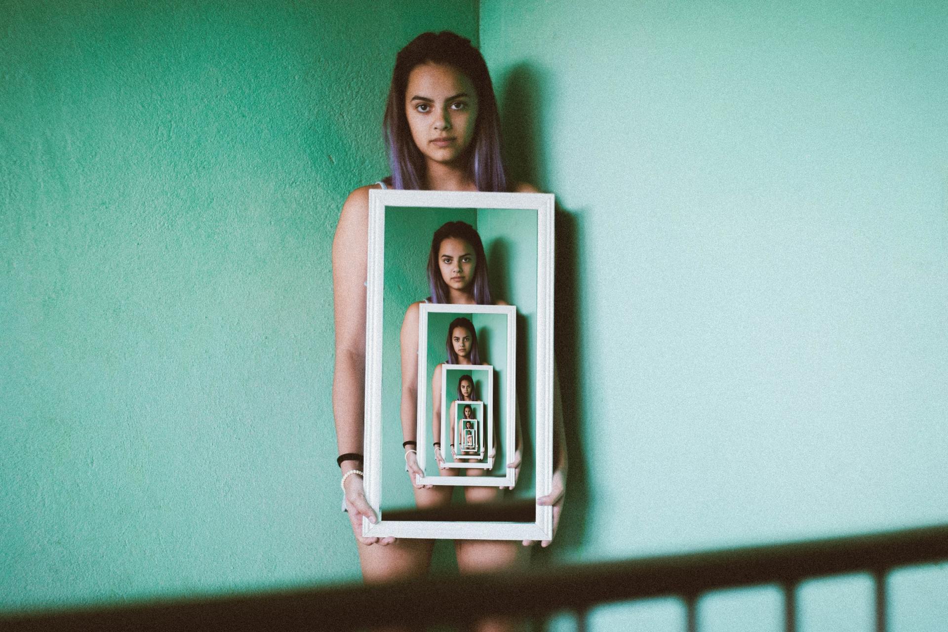 Woman holds a mirror with her reflection infinitely repeated, standing in a turquoise corner.
