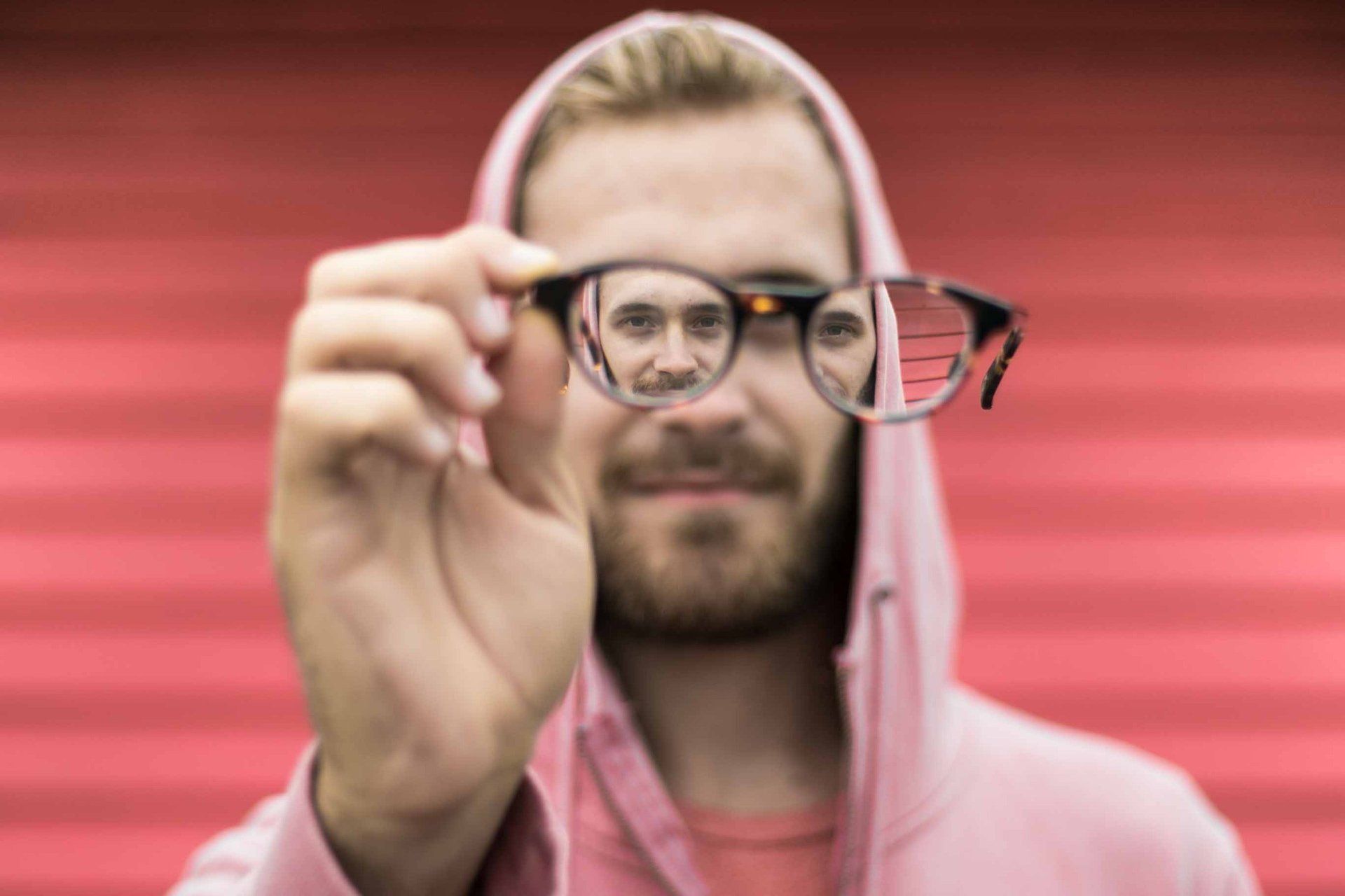 Mann mit Brille, in der sich sein Gesicht spiegelt, trägt einen rosa Kapuzenpullover vor rotem Hintergrund.