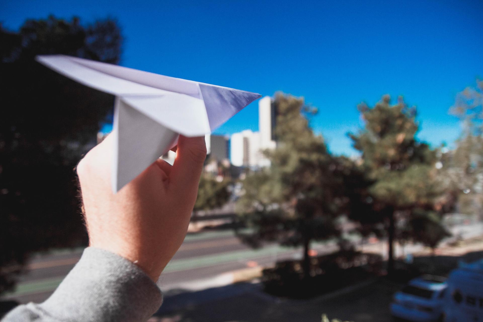 Mão segurando um avião de papel na direção de uma rua parcialmente vazia. É possível ver árvores no meio e nas laterais da rua e o céu está azul e sem nuvens. Dois carros aparecem no canto da foto.