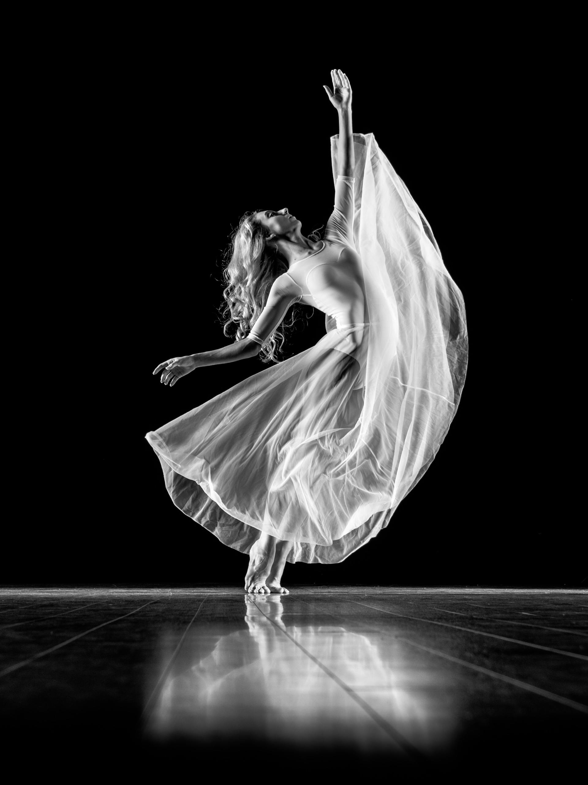 Ballerina in white flowing dress, leaping on a stage with arms raised, black backdrop.