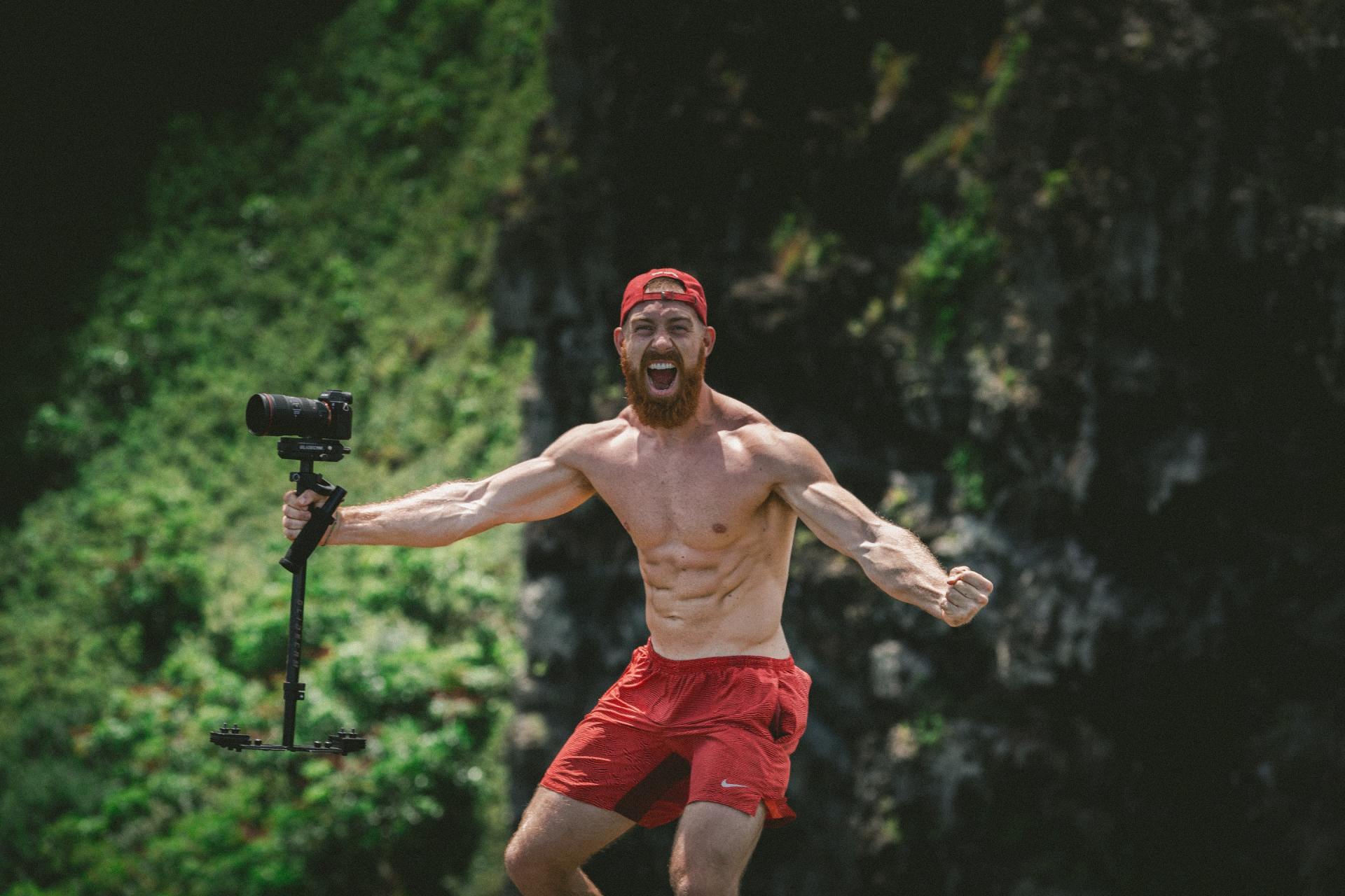 Shirtless man in red shorts and hat yells, holding camera, standing on a rock in front of green foliage.