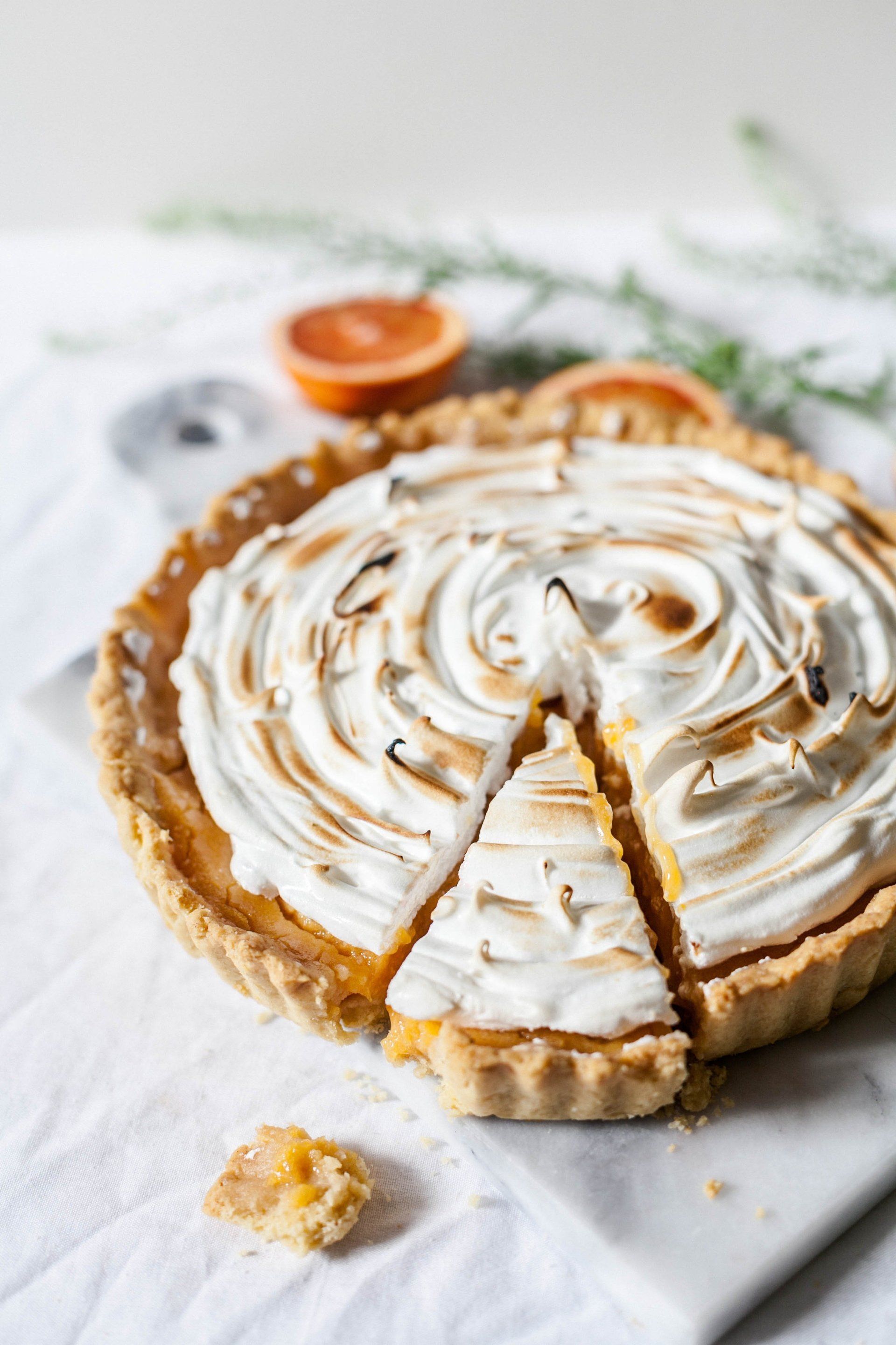 Lemon meringue tart on a white surface, slice removed, surrounded by citrus and greenery.