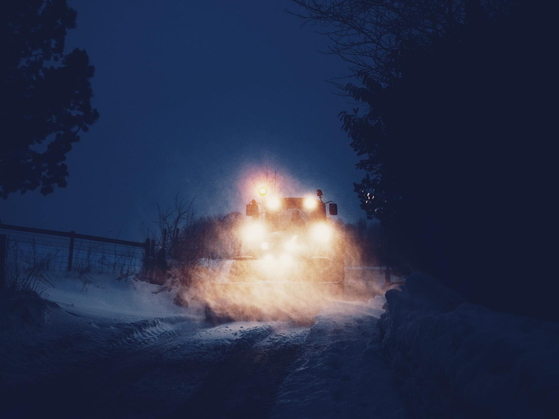Tractor with bright headlights driving through snow at night on a rural road.