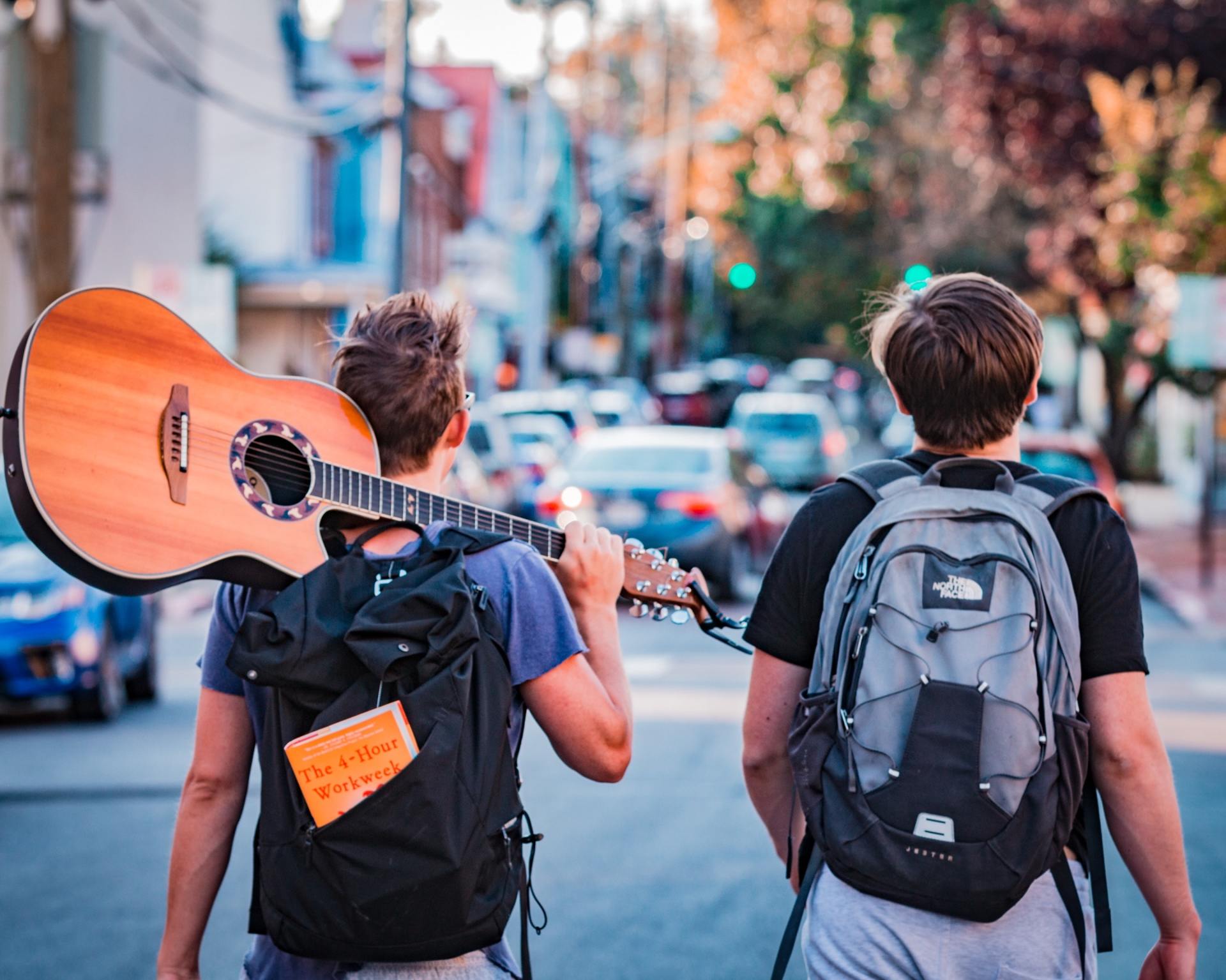 Two men with backpacks are walking down a street one is carrying a guitar