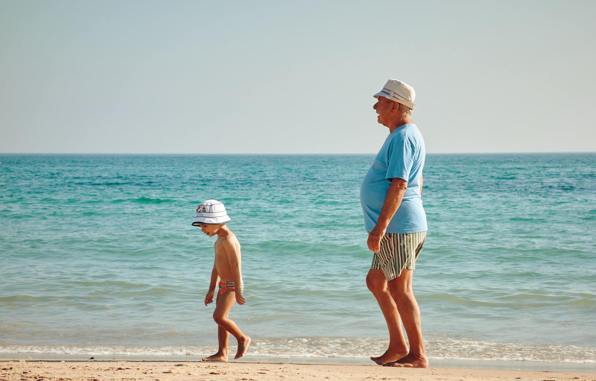 Older man probably grandfather walking on oceanside beach with younger boy probably grandson.