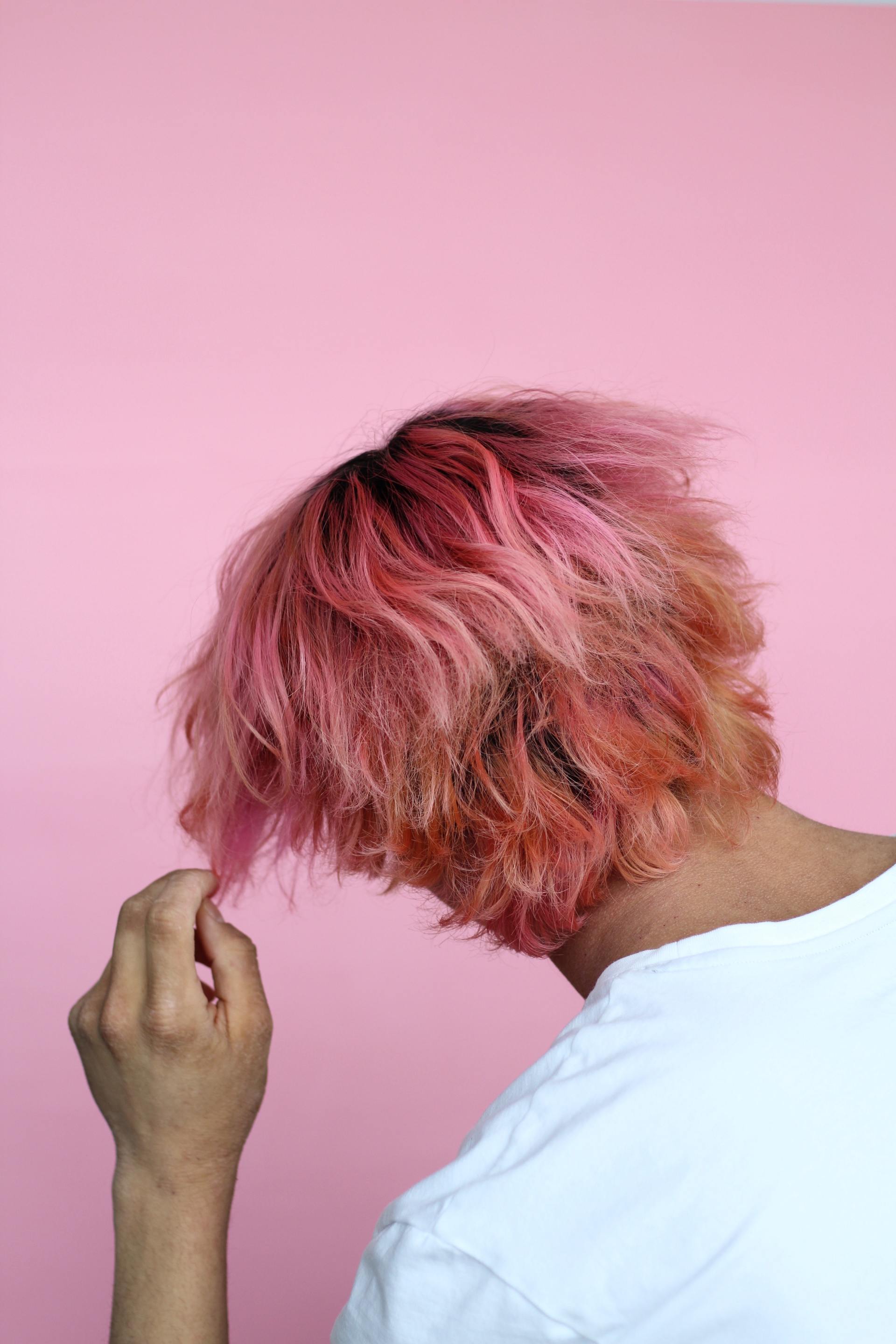 A person with pink hair is holding their hair in front of a pink background.