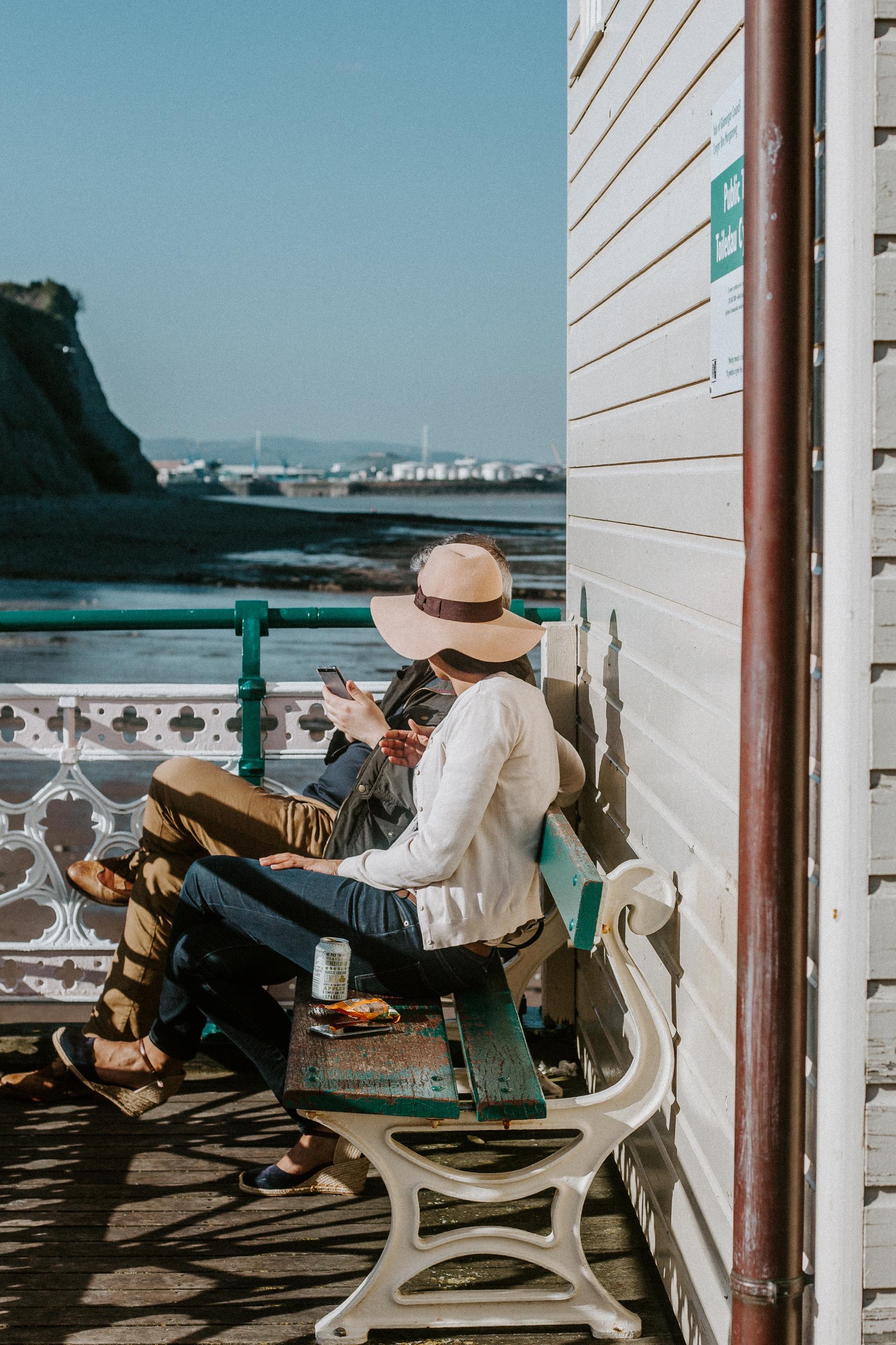 A woman in a hat is sitting on a bench looking at her phone.
