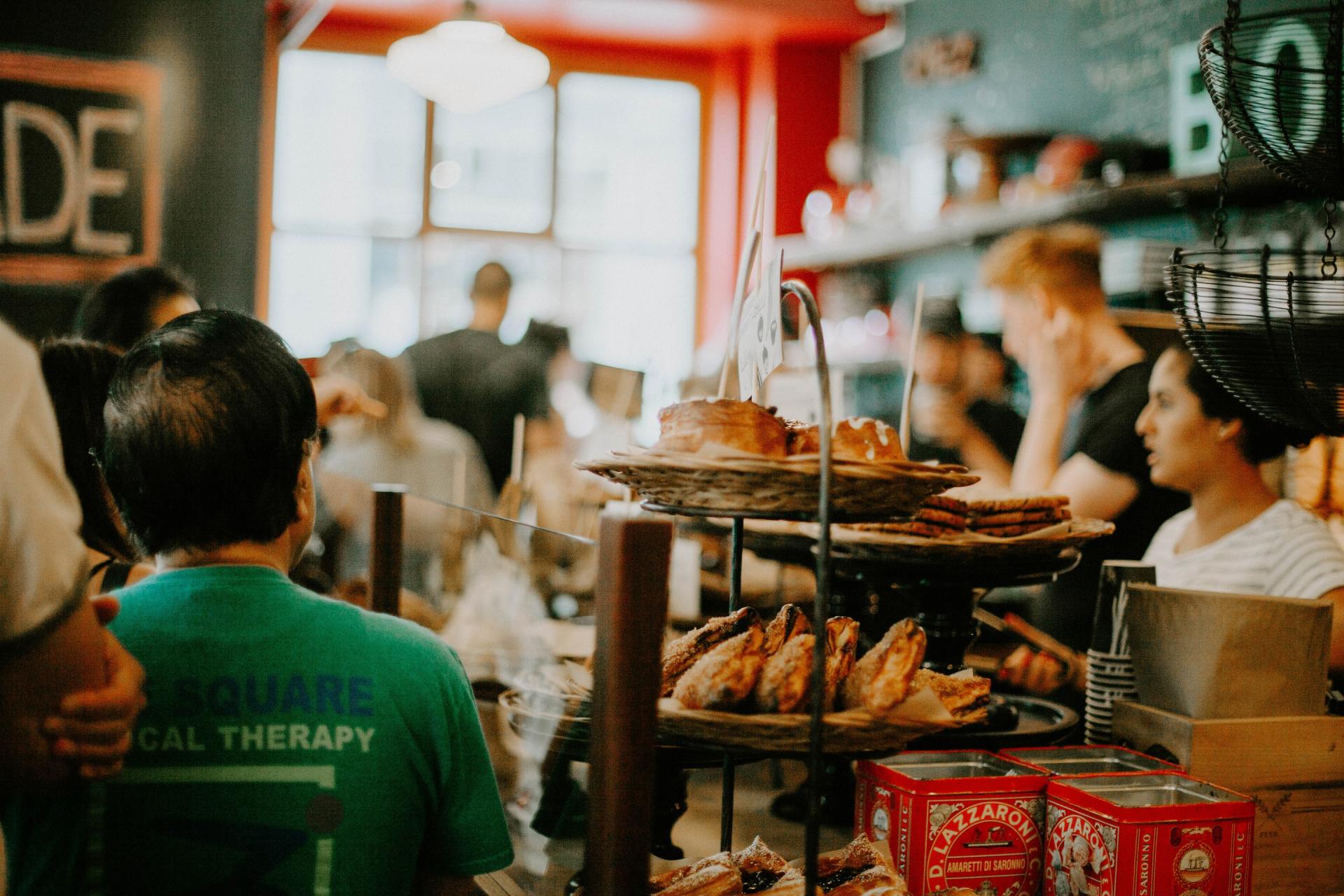A crowded bakery. People in line, pastries on display. Green shirt, red walls, warm lighting.