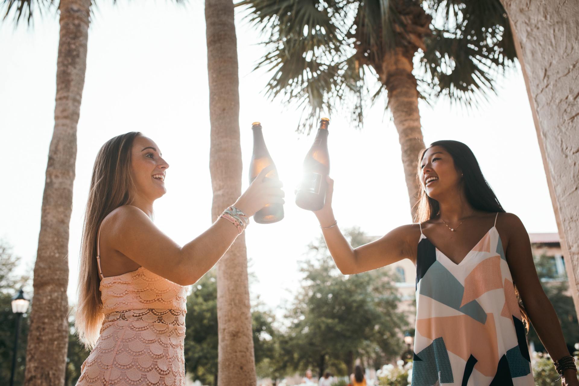 Two women are toasting with champagne bottles in front of palm trees.