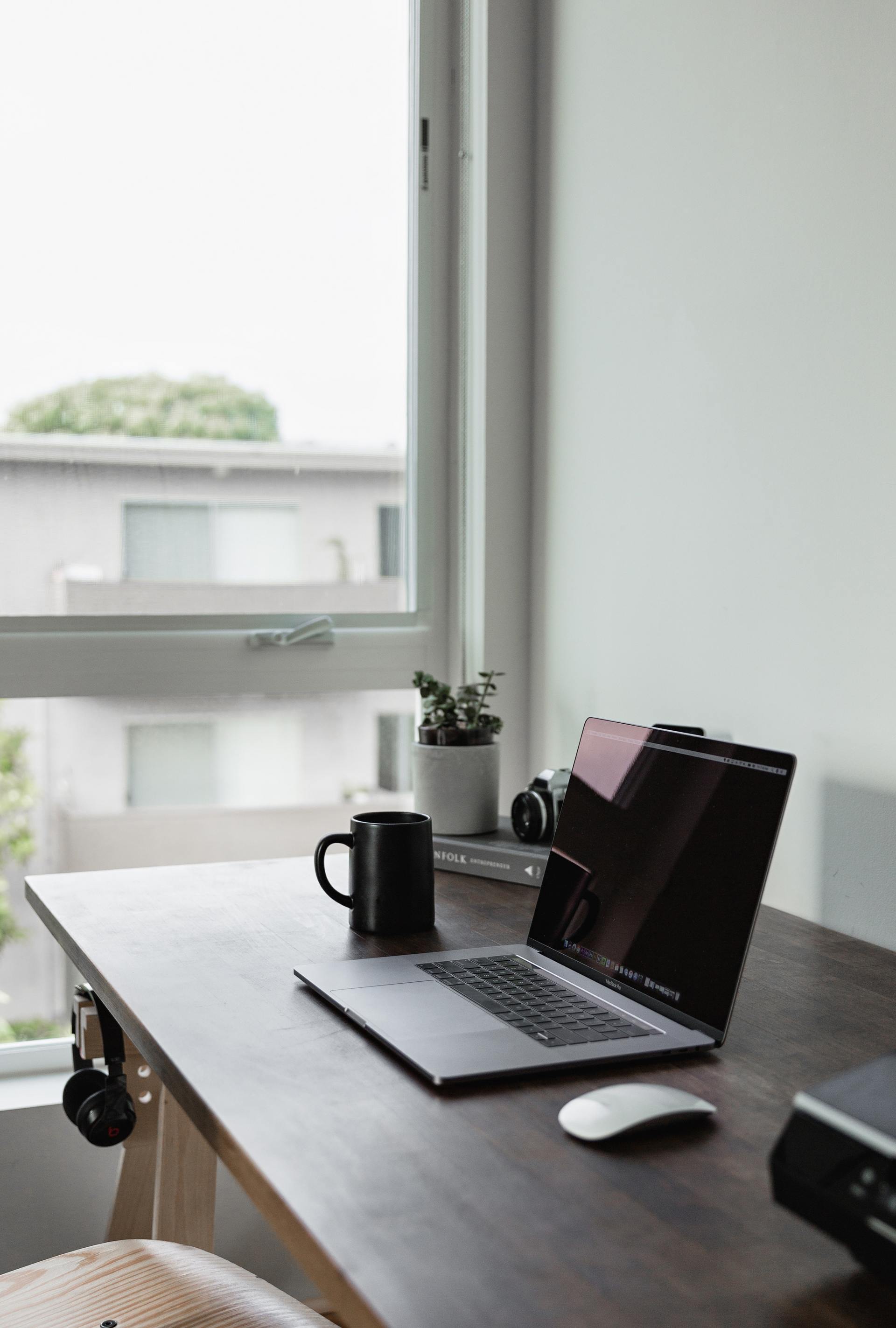 Laptop, mug, and plant on a wooden desk by a window; workspace with a camera and headphones.