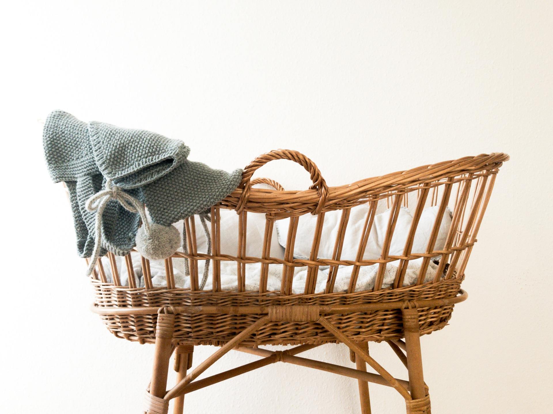 A photo of a wicker bassinet with a gray knit blanket draped over the left side, set against a light background.