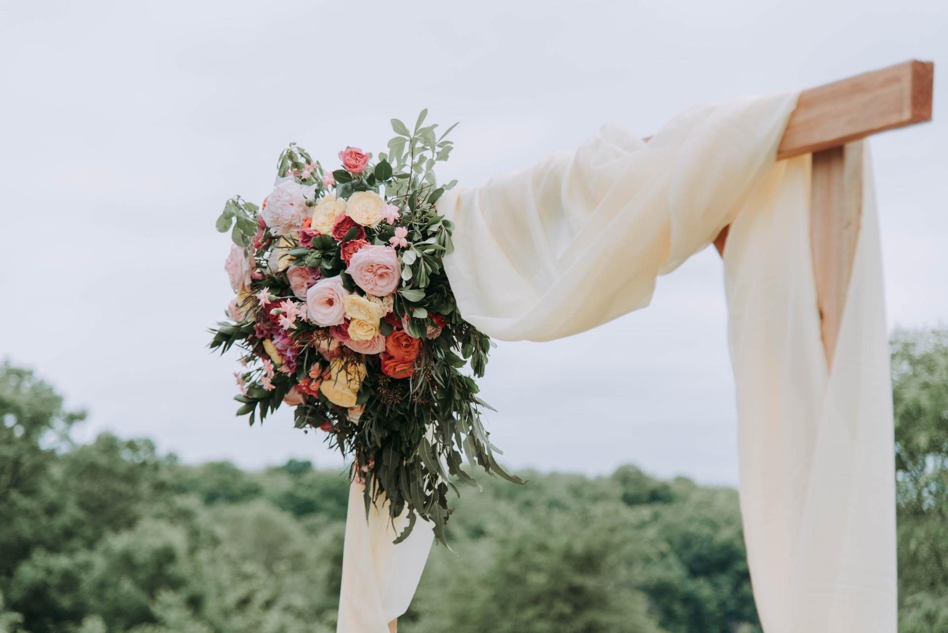 A photo of the top of a square wooden archway with white fabric draped over it and a large bouquet of flowers on the lefthand side