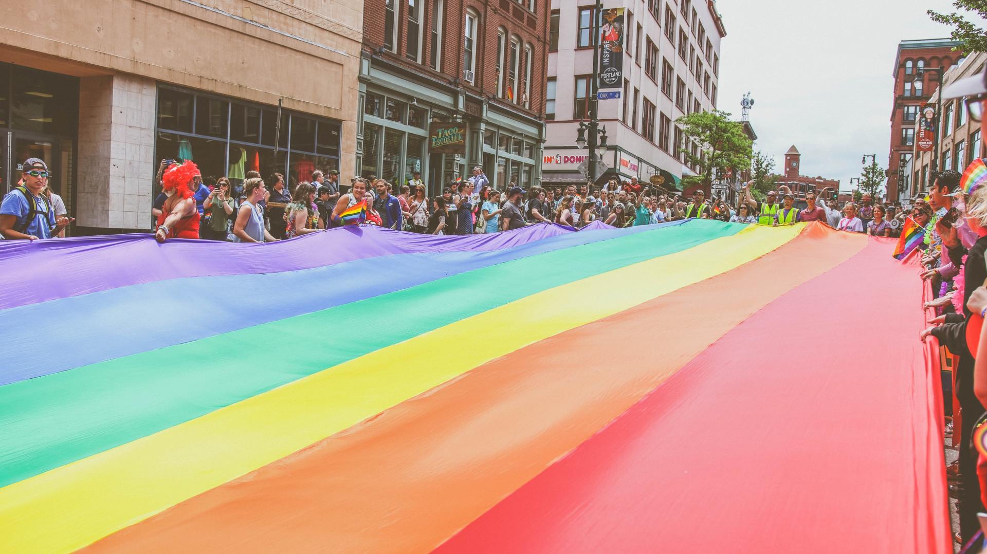 A photo of a long rainbow flag being stretched across a street during a Pride parade on Congress Street in Portland