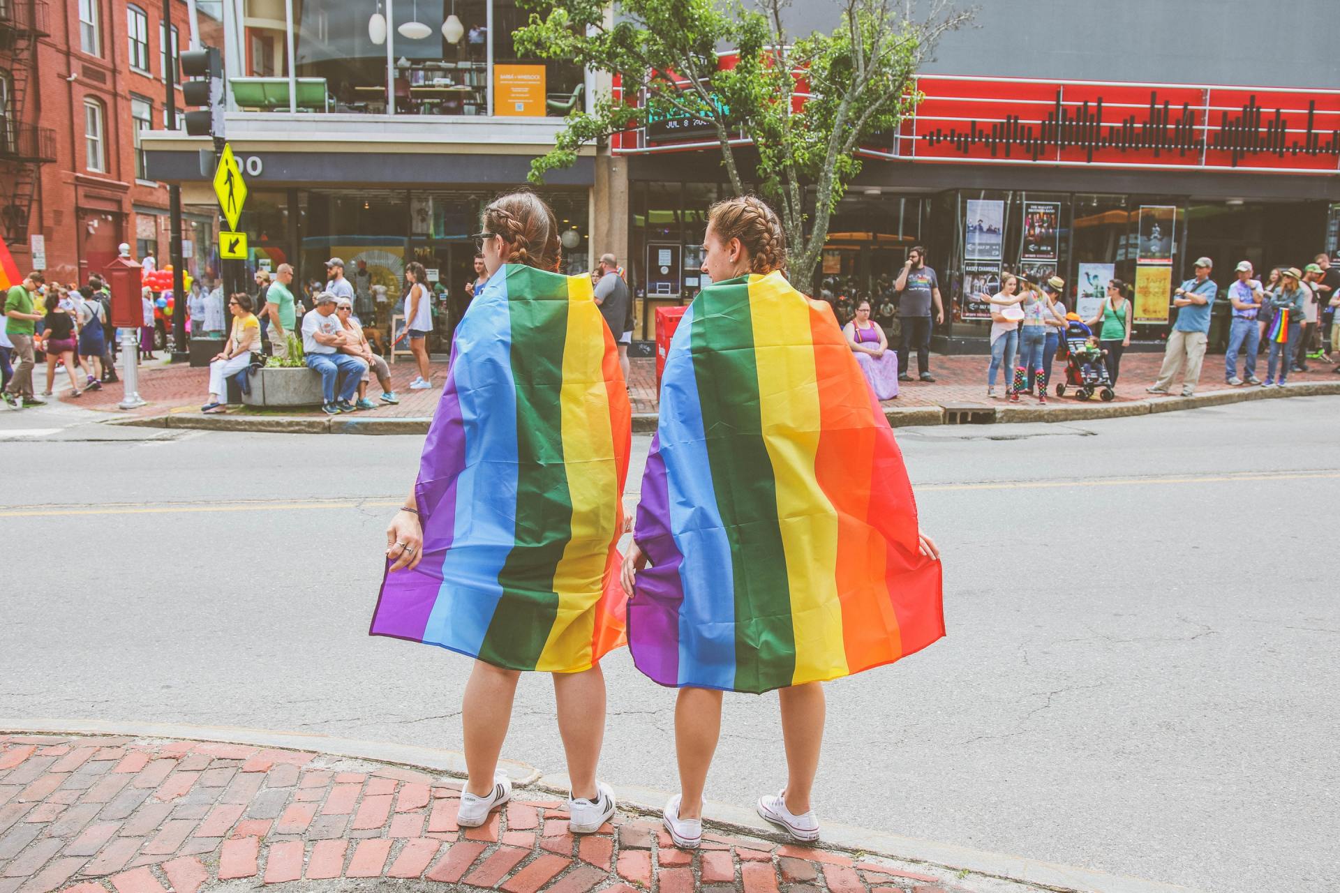 Two people are standing on the side of the road holding rainbow flags.