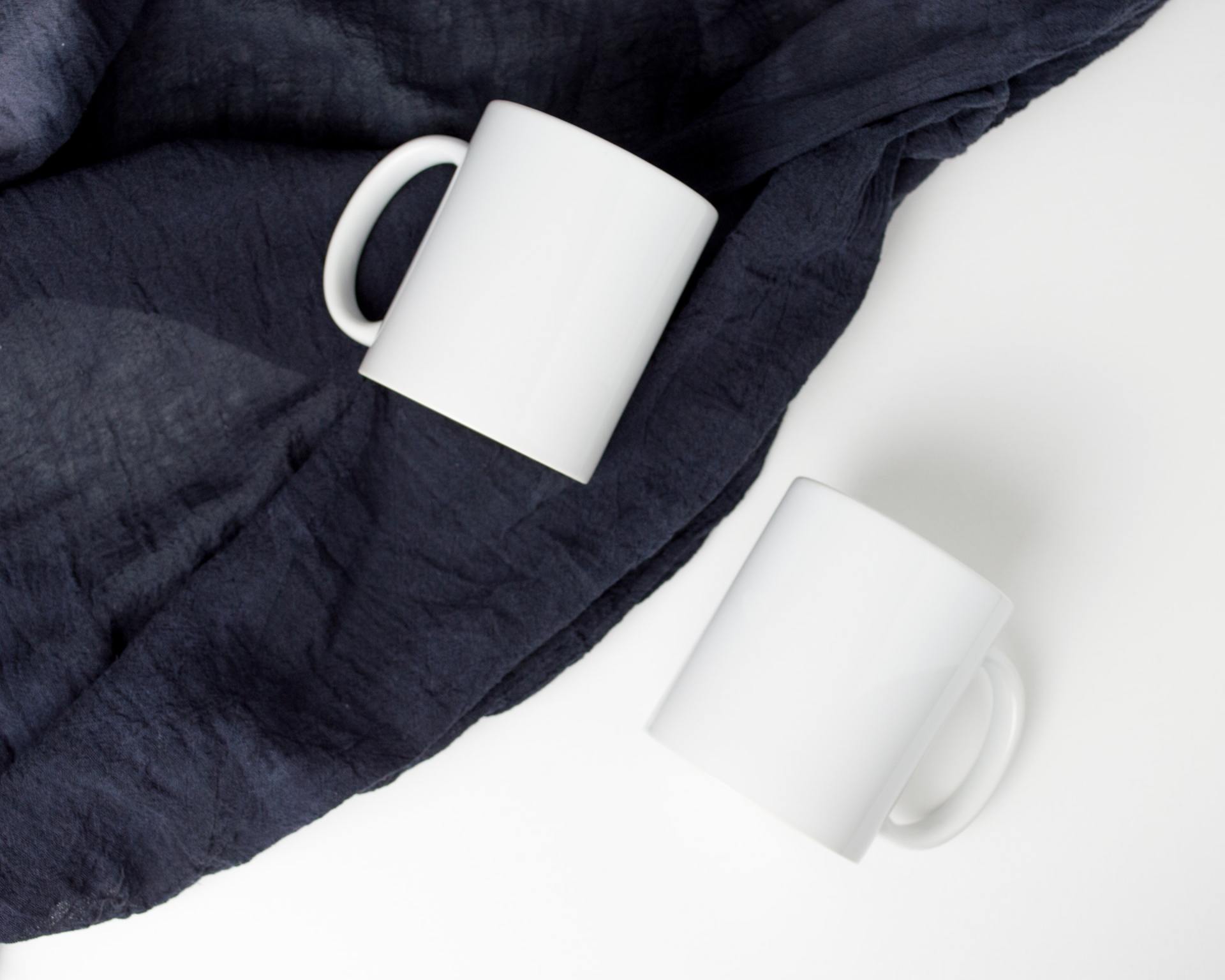 Two white ceramic mugs on their side with blue linen drapped around it