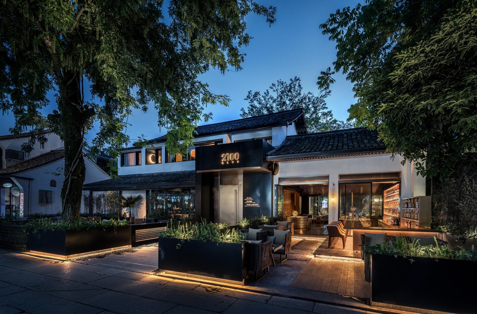 Restaurant exterior at dusk, illuminated by lights; large black planters line the entrance, trees overhead.