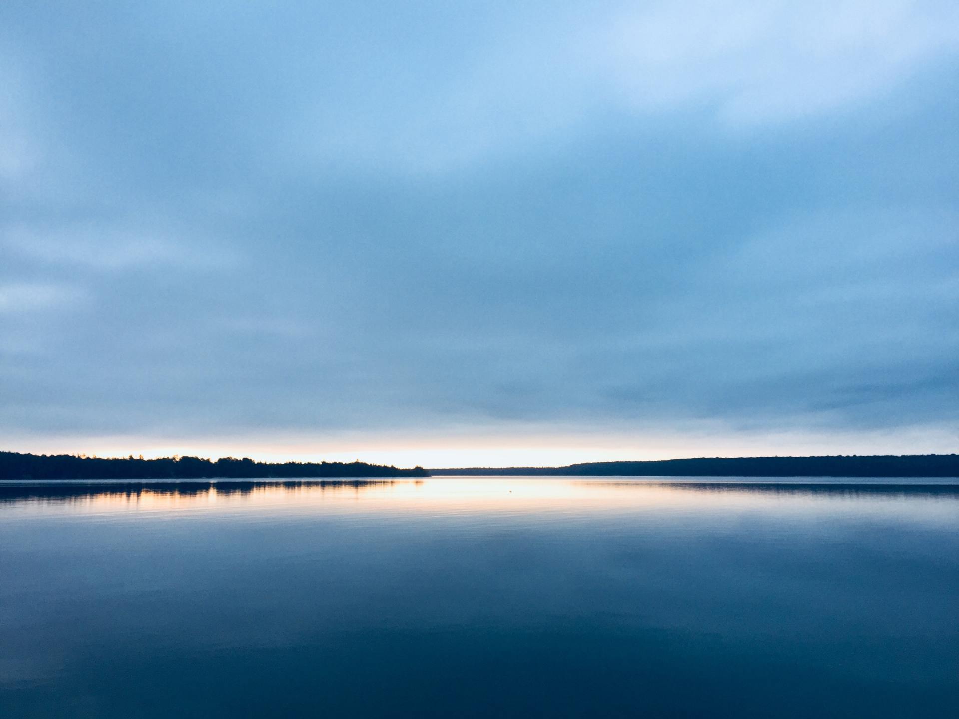 Calm lake reflecting overcast sky with a hint of sunrise. Silhouetted trees line the horizon.