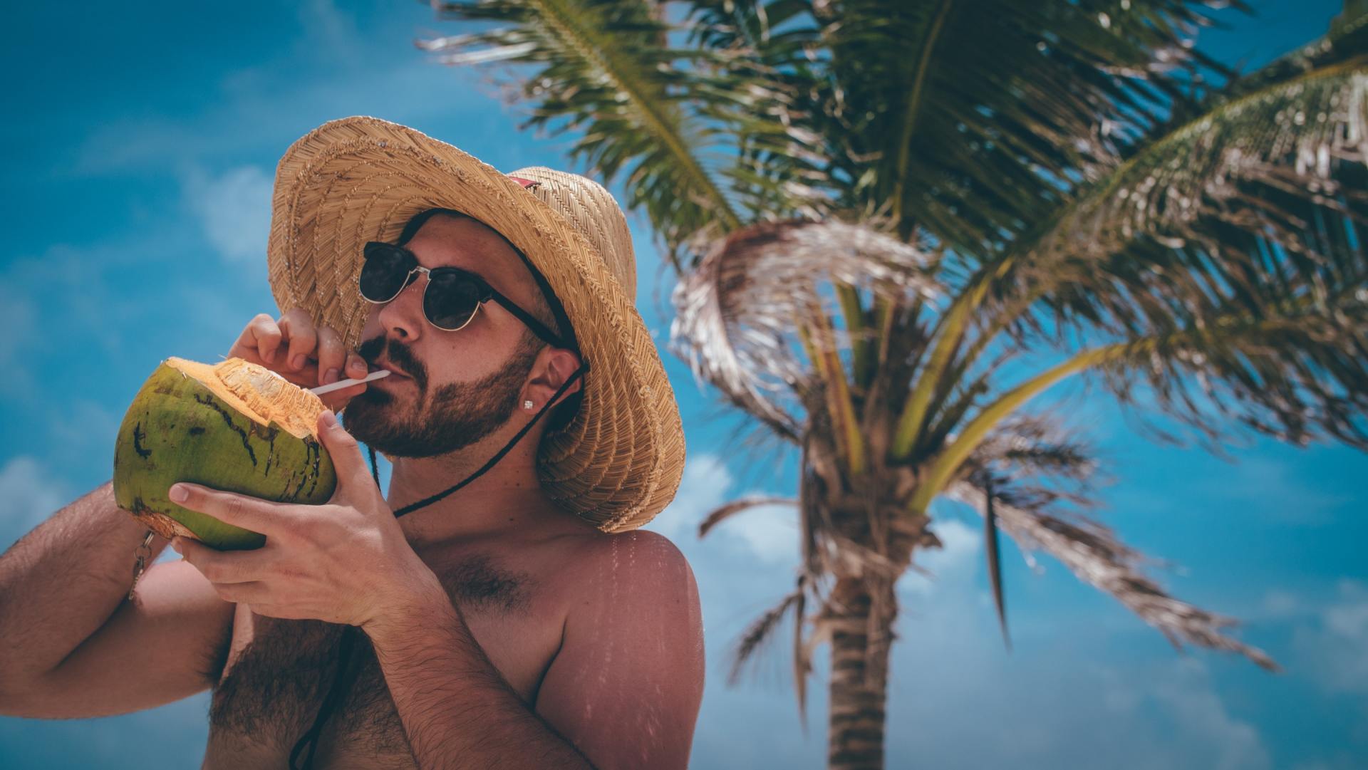 man with sunglasses and a straw hat drinking a coconut in front of a palm tree