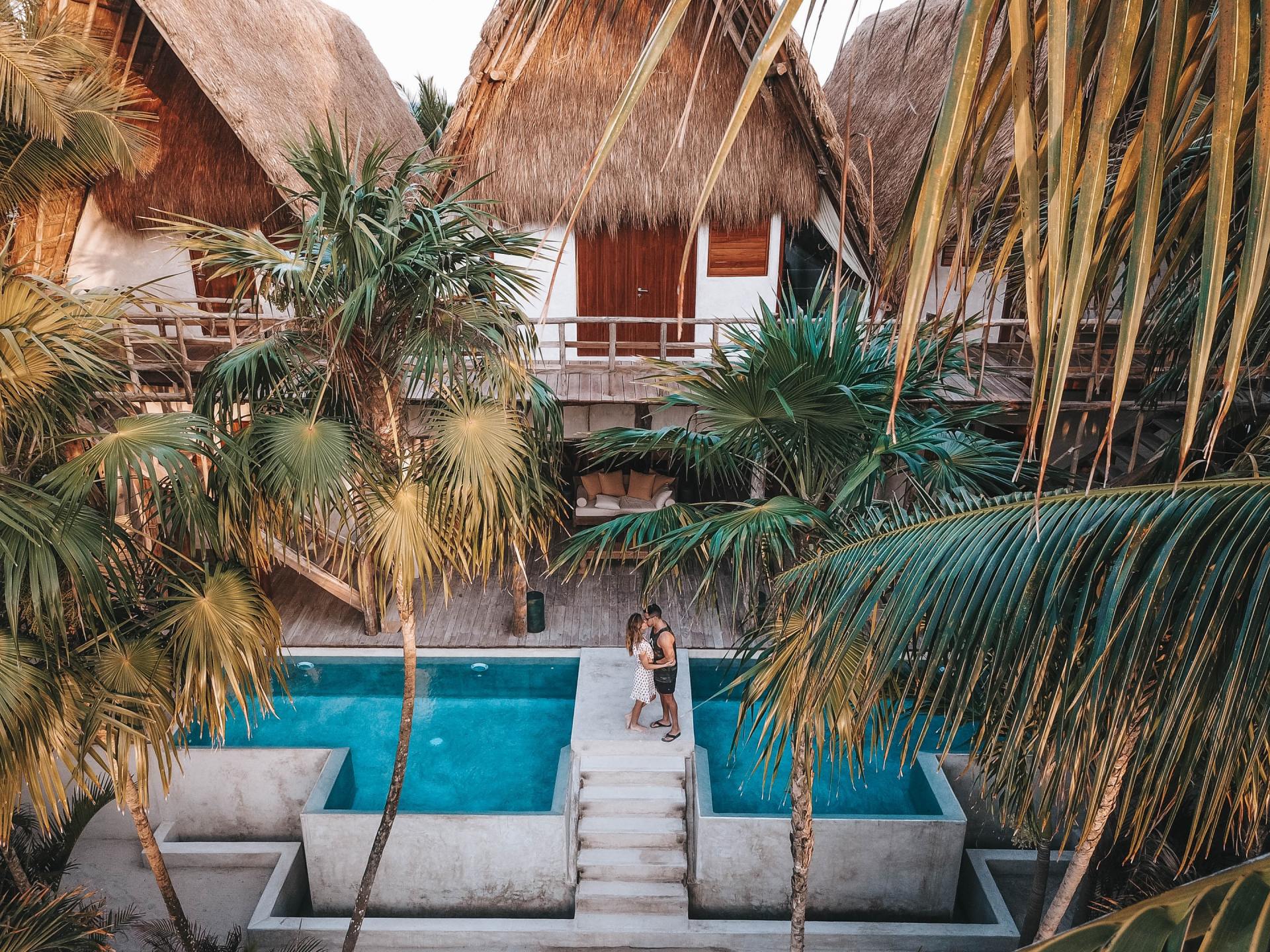 A couple is standing next to a swimming pool in front of a house surrounded by palm trees.