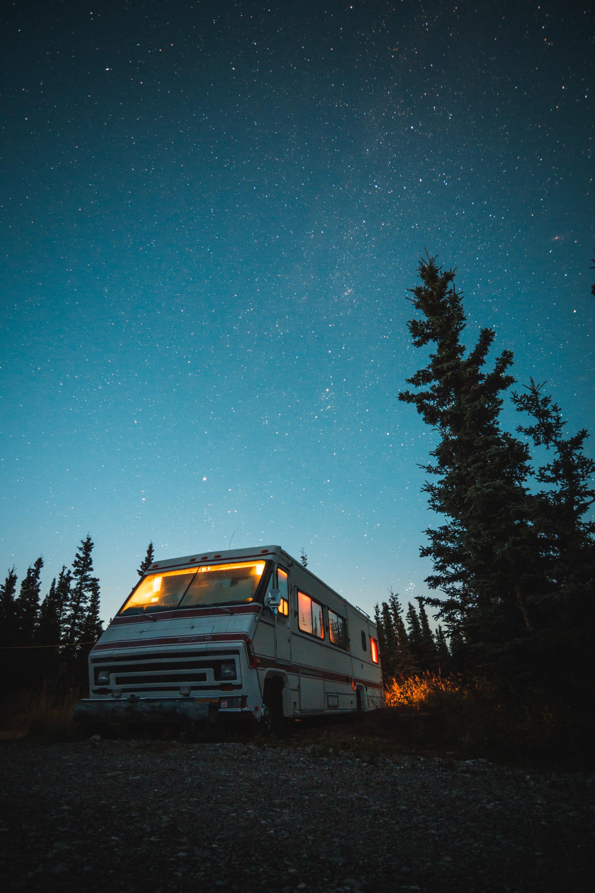 RV parked under a starry night sky, with warm interior lights glowing, and trees surrounding.