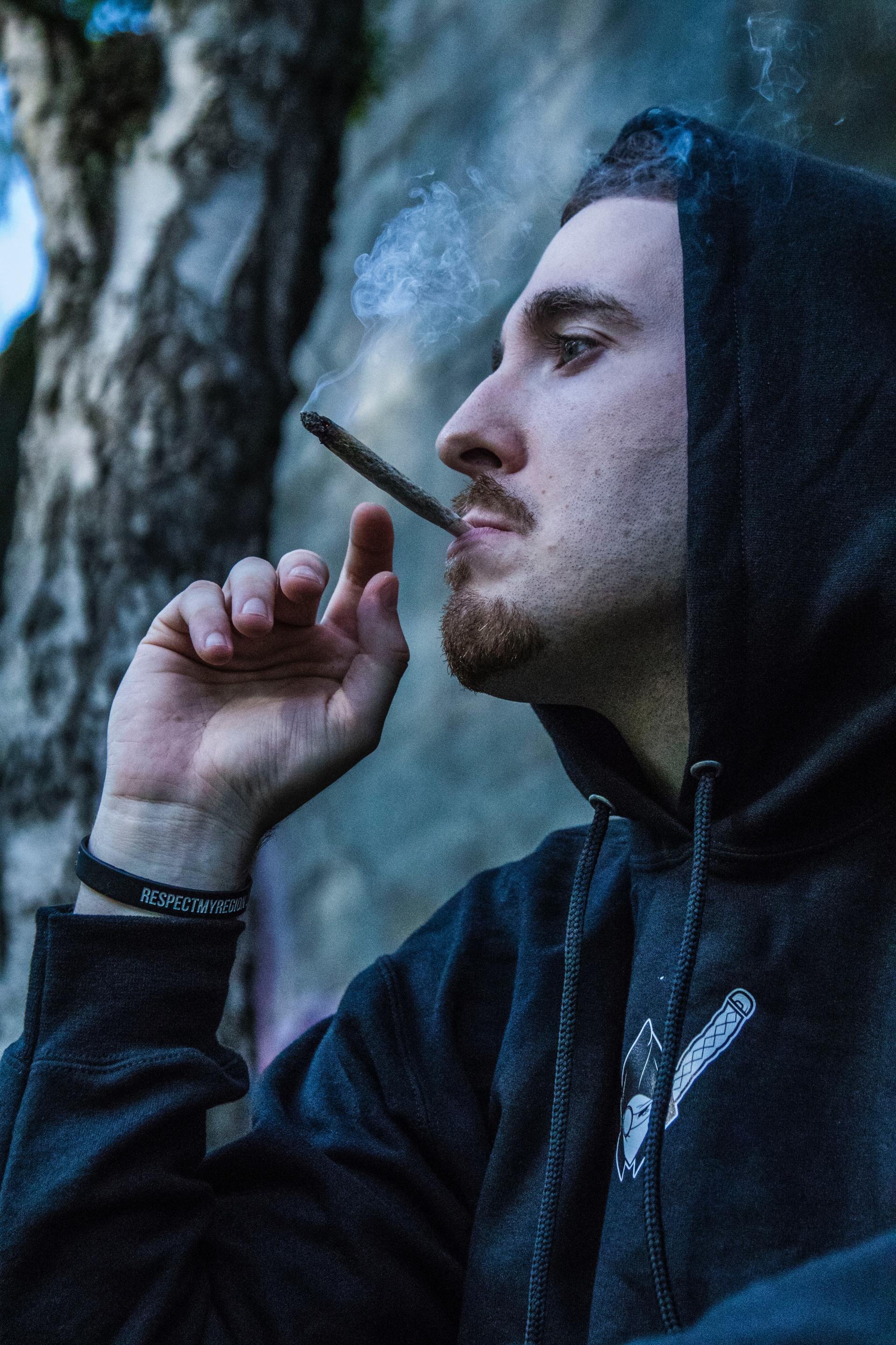 Man in a black hoodie smokes a cigarette, exhaling smoke. He's outside near a tree, looking to the side.