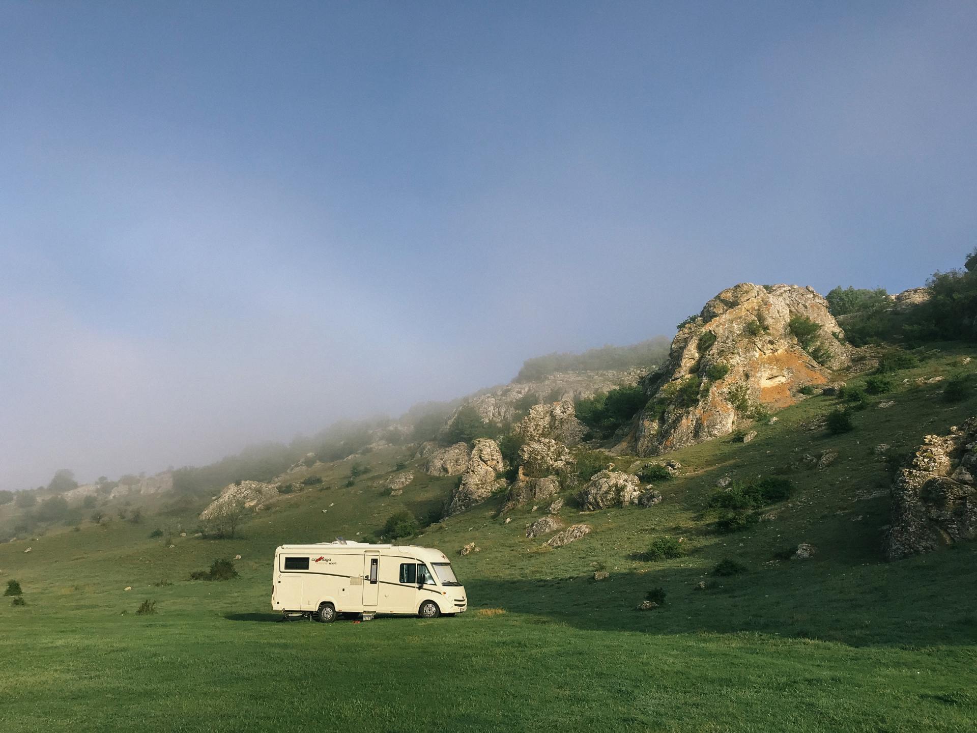 Camper van parked on grassy field with rocky hills and a misty sky in the background.