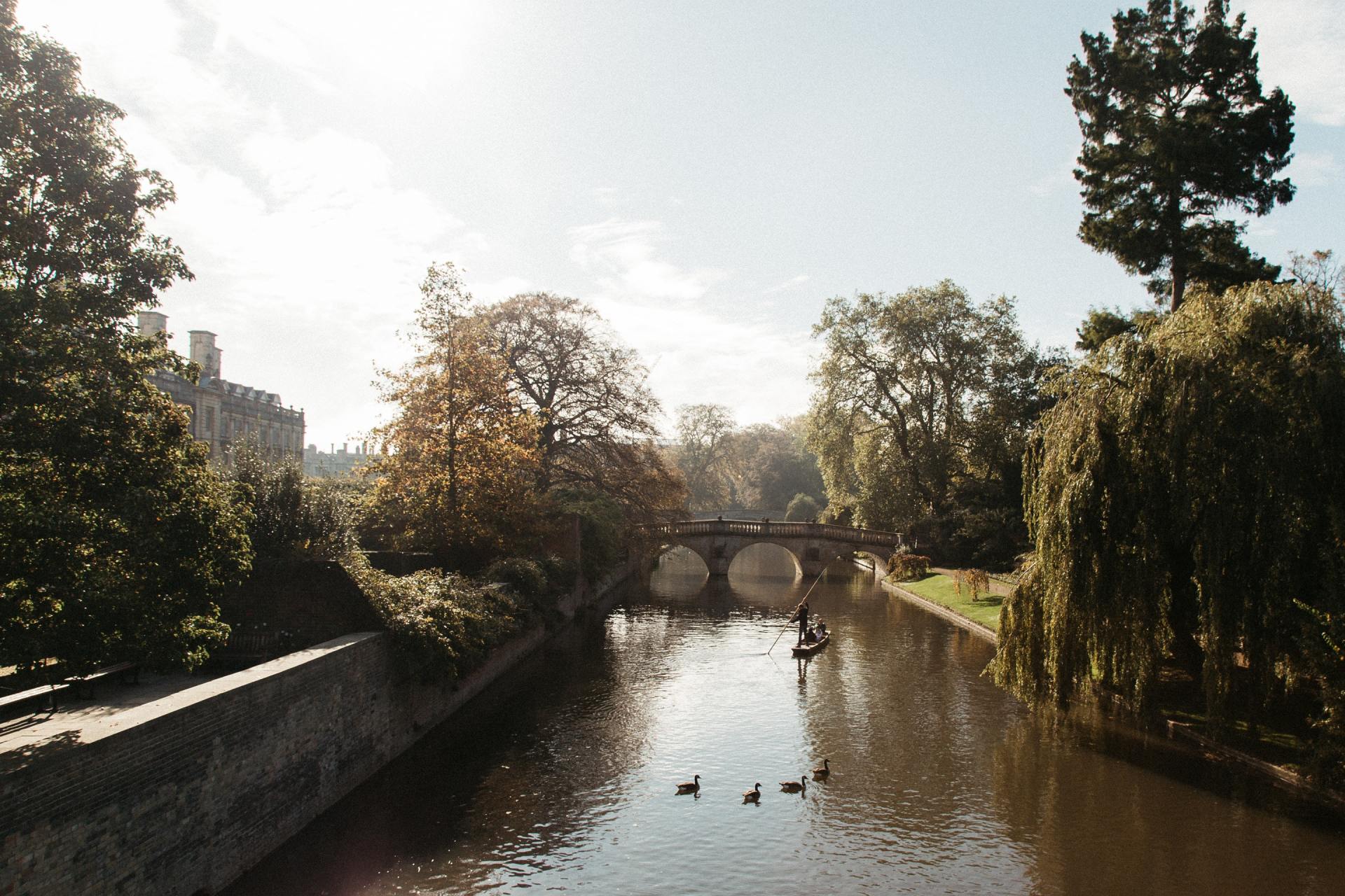 Hazy afternoon photo of river and bridge in Cambridge. Punt boat in middle-distance. Trees either side.