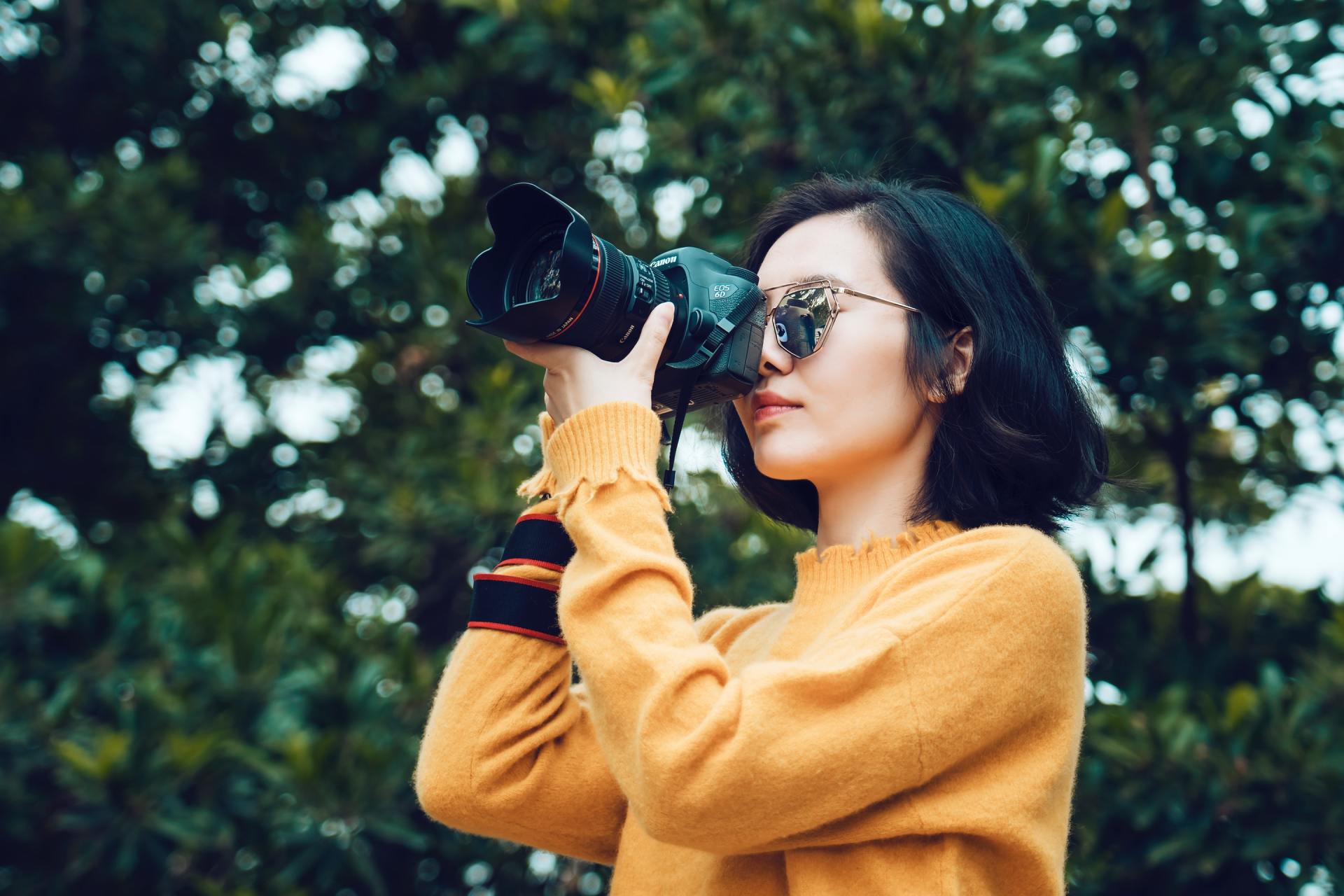 A woman in a yellow sweater is taking a picture with a camera.