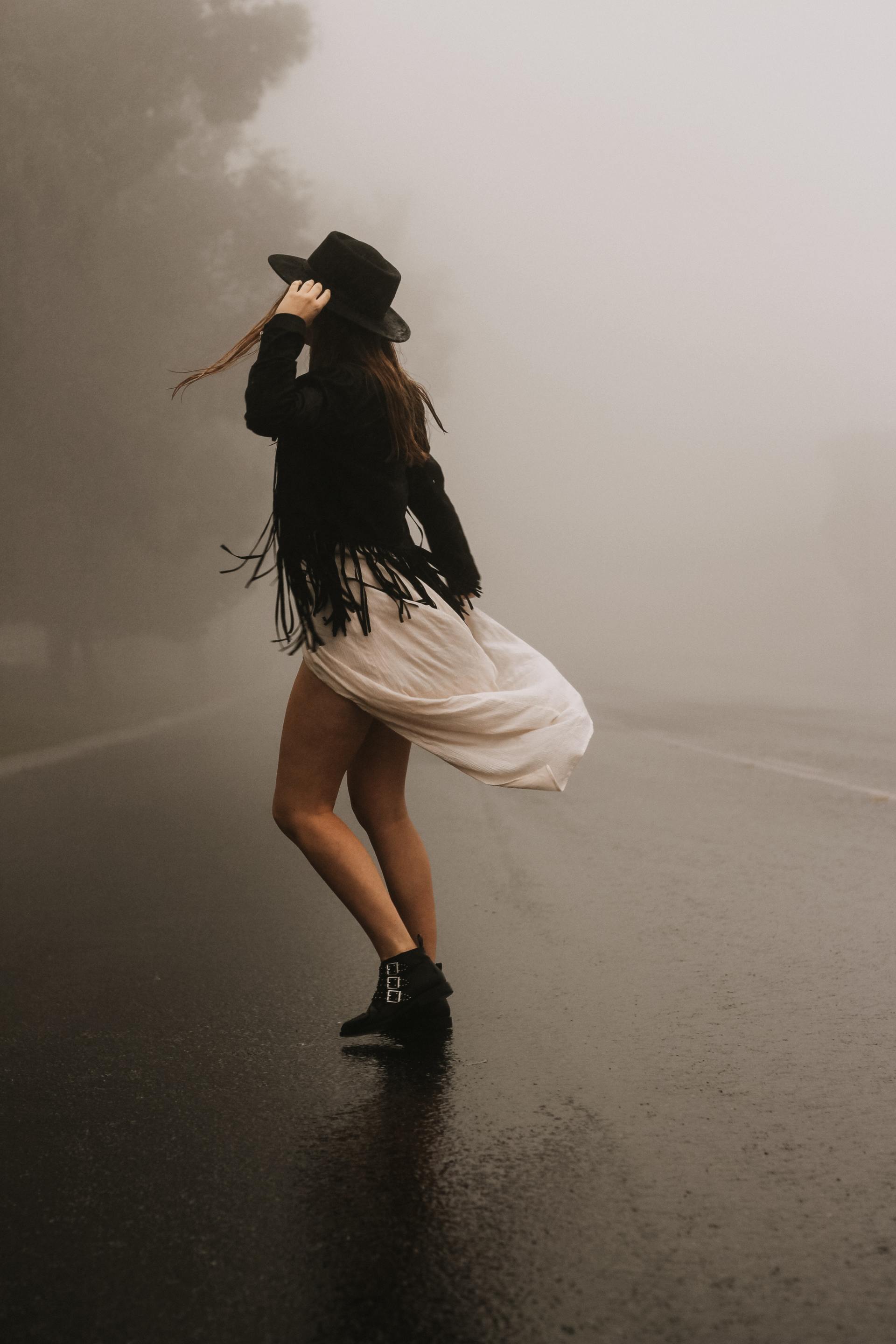 A woman in a white dress and black hat is standing on a foggy road.