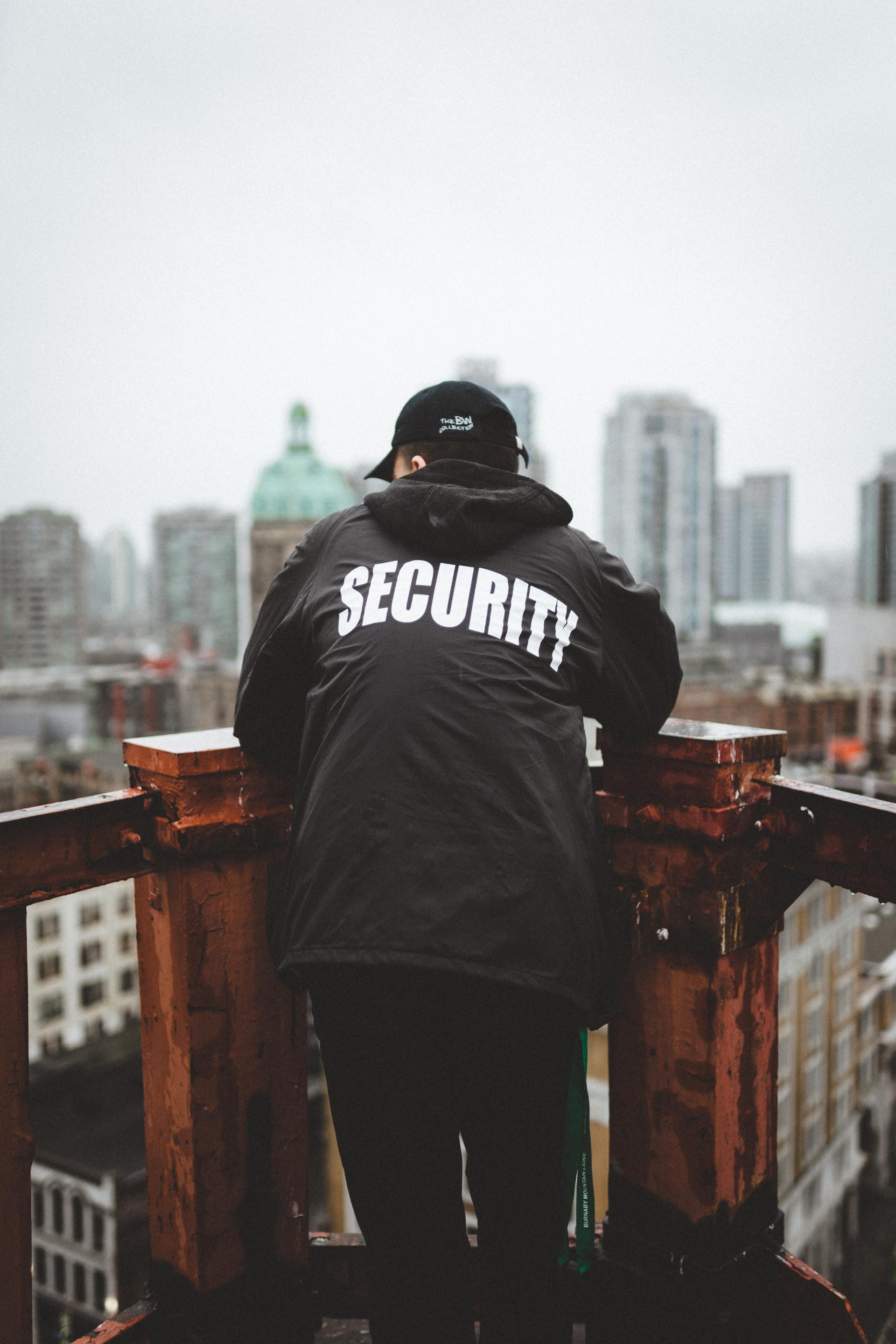 Security guard on a rooftop overlooking city, wearing black jacket with
