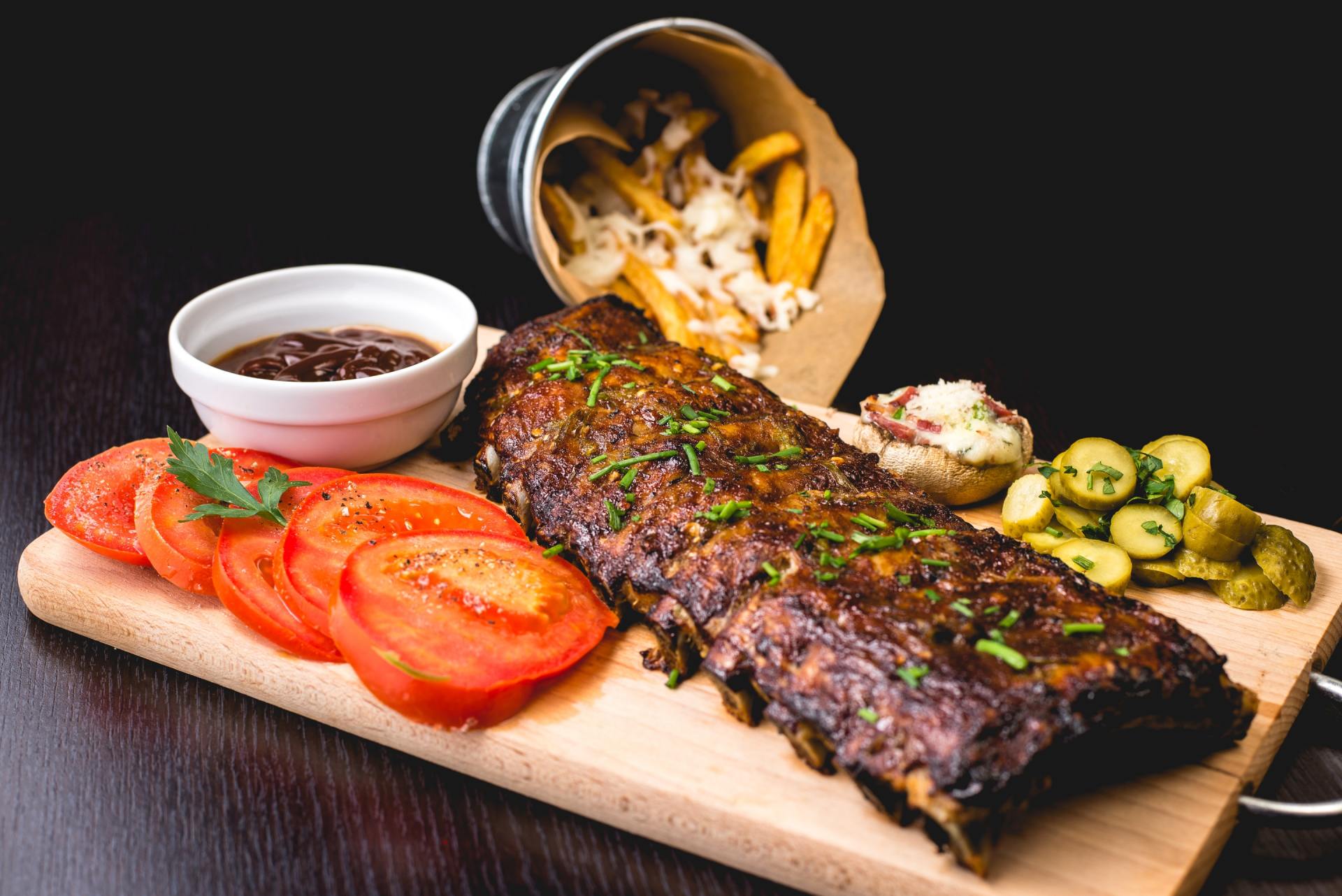 A wooden cutting board topped with ribs , tomatoes , pickles and french fries.