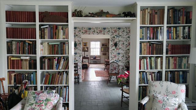 Built-in bookshelves flank a doorway, leading to a floral wallpapered room. Comfortable armchairs, plants, and books abound.