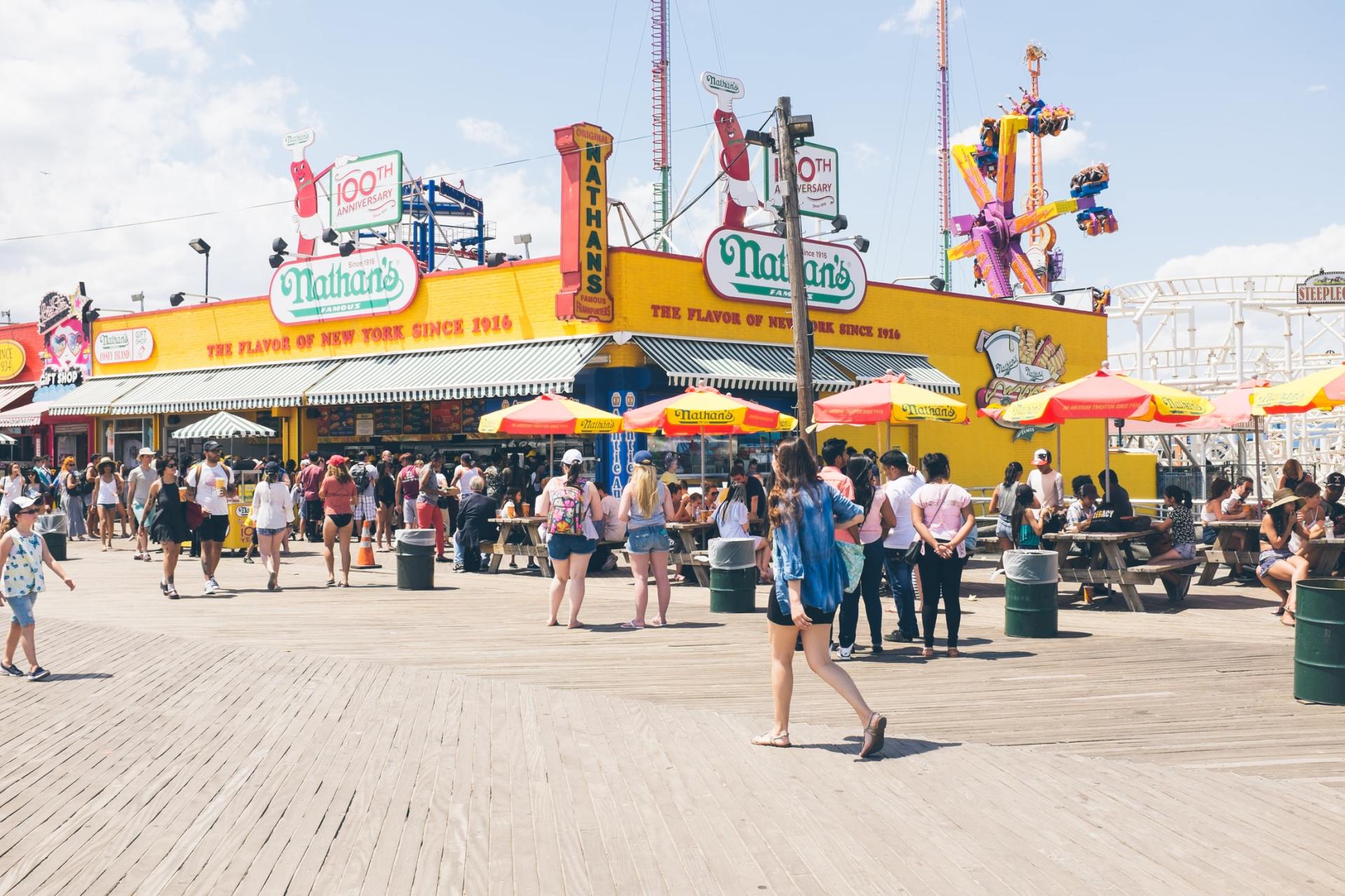 A group of people are walking on a boardwalk in front of a hot dog stand.