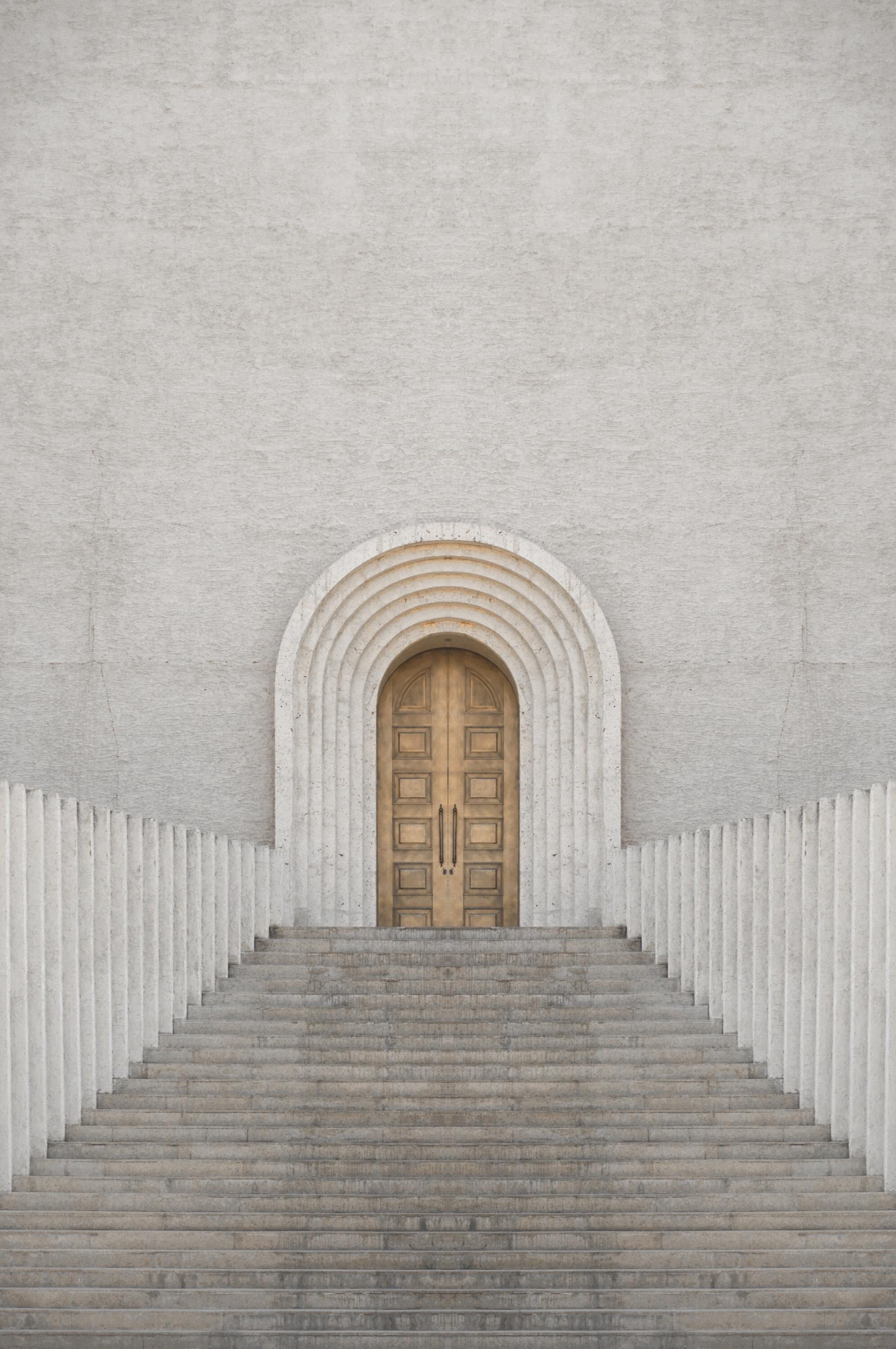 Steps leading up to a light-brown door set in a light-gray, arched doorway within a textured, tall building facade.