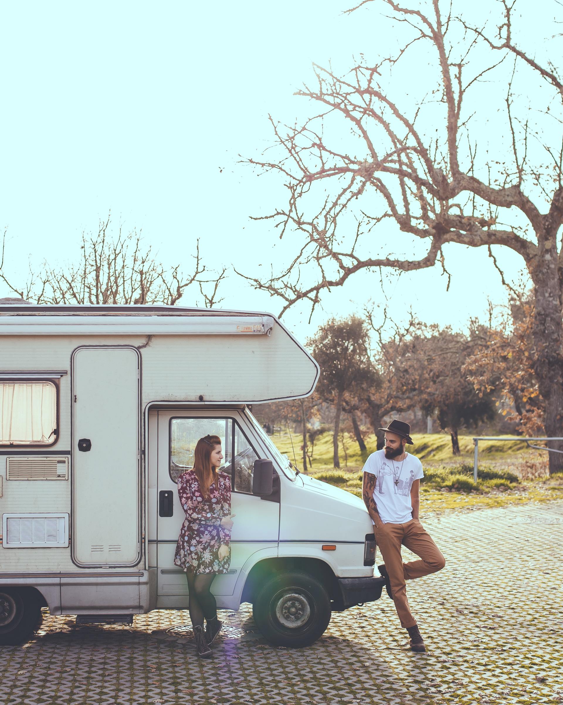 Couple by a white campervan, sunny outdoors. Woman leans, man poses, tree backdrop.