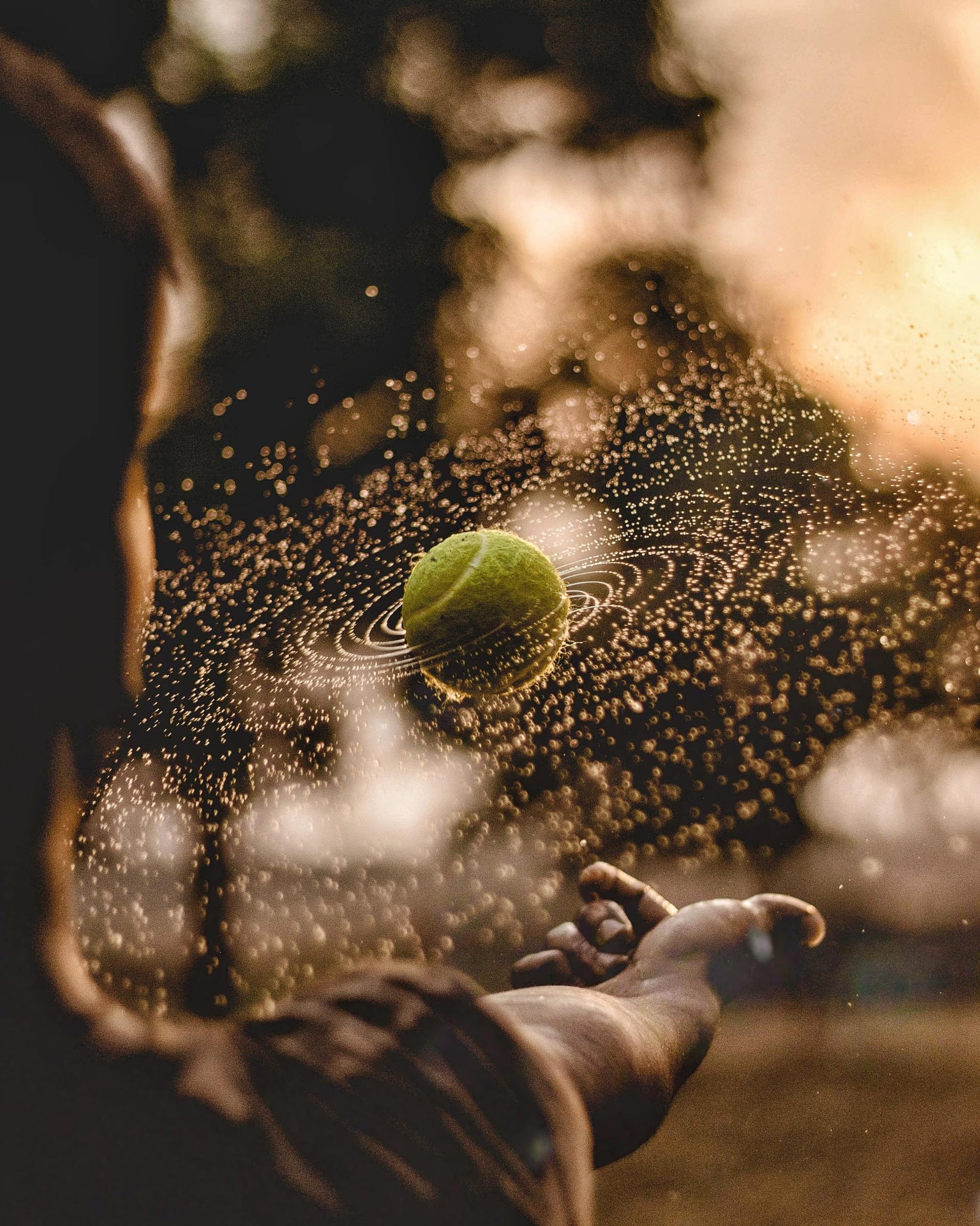 Person throwing a tennis ball, with water droplets and golden light.
