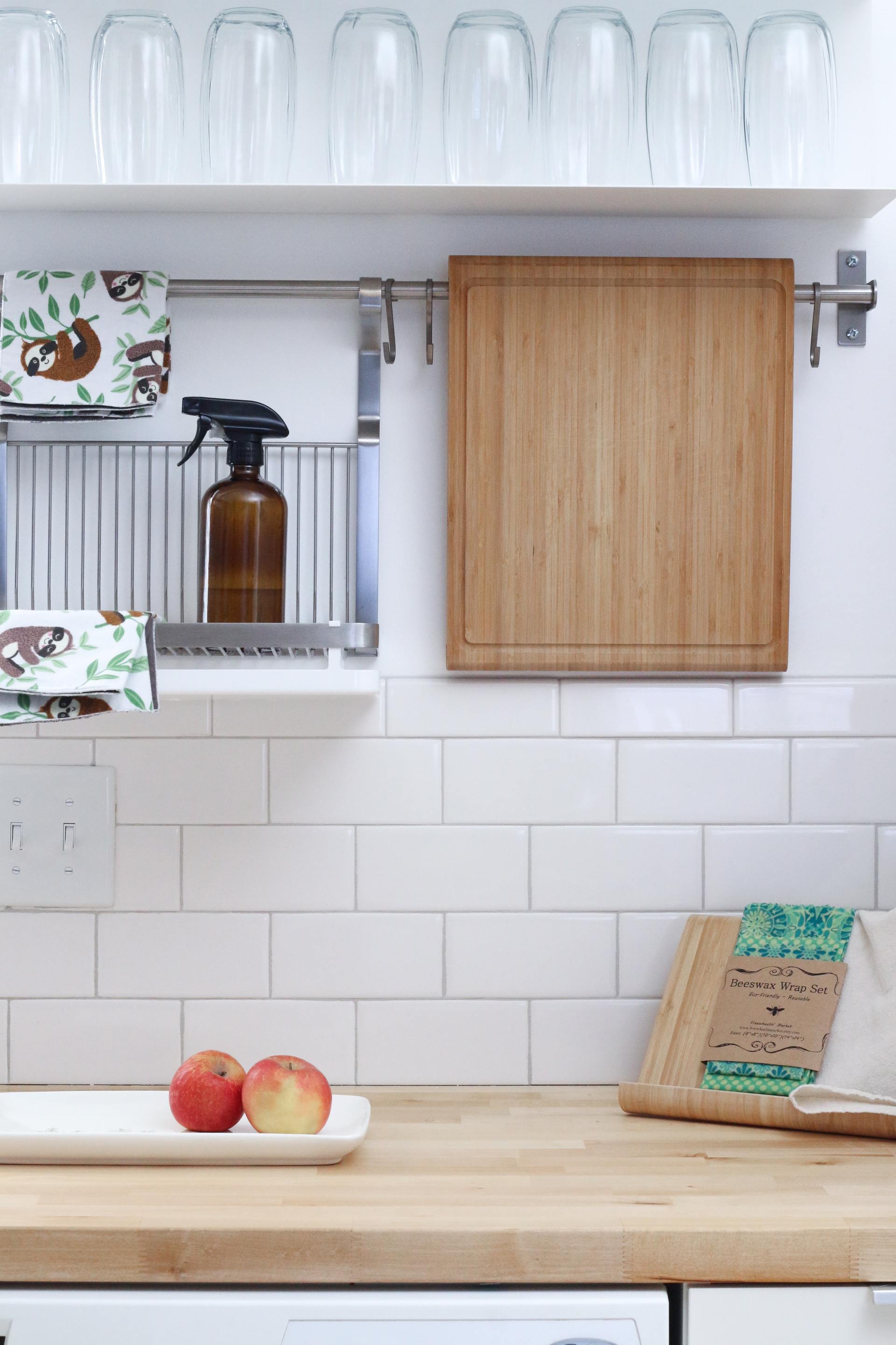 Image depicts a kitchen are with white tiles  chopping board on the bench and some cleaning products.